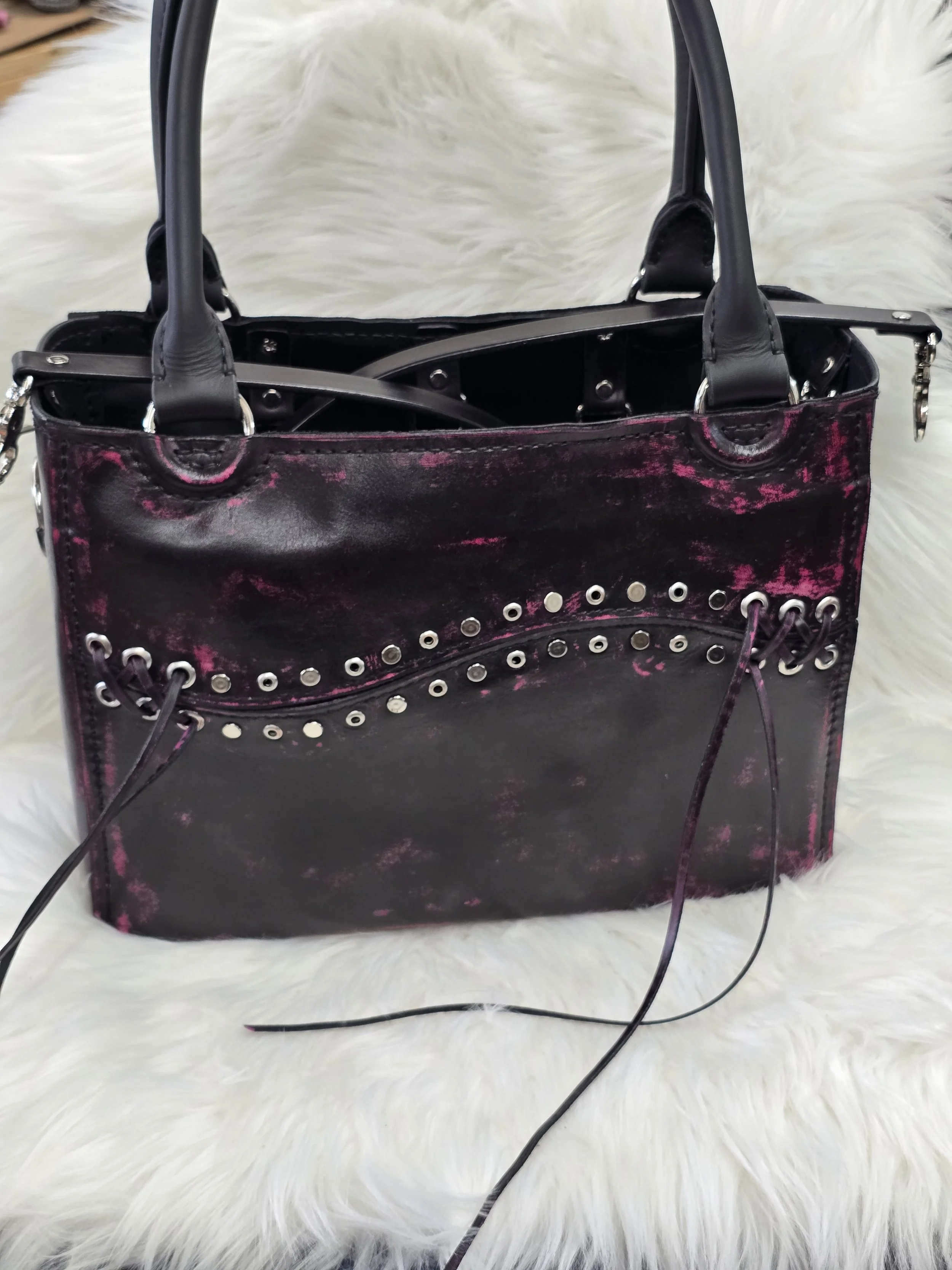 Black and pink distressed leather handbag with metal eyelets and a shoulder strap, placed on a white furry surface.