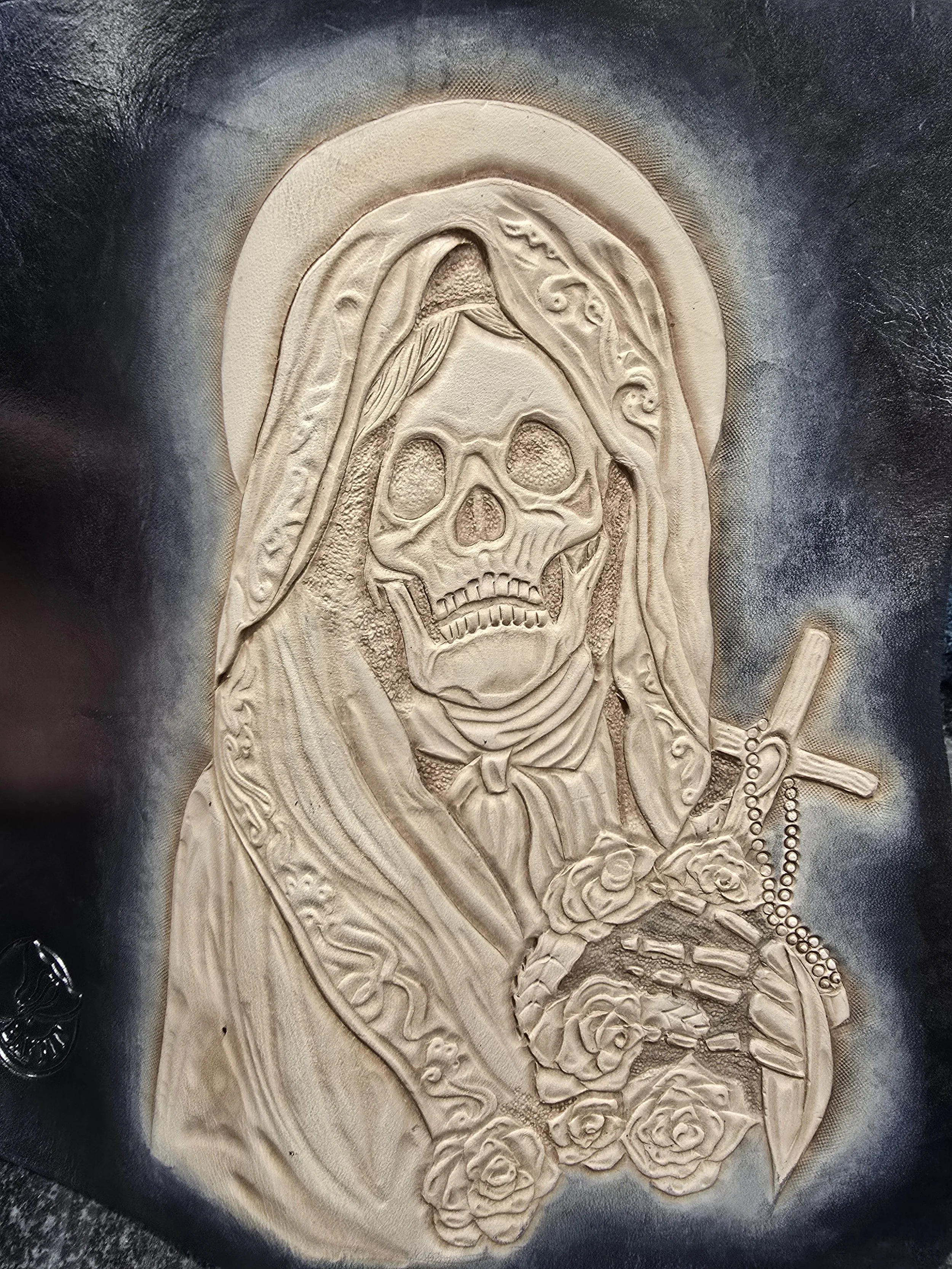 A carved relief of a skull with long hair, holding a cross and rosary, surrounded by roses.