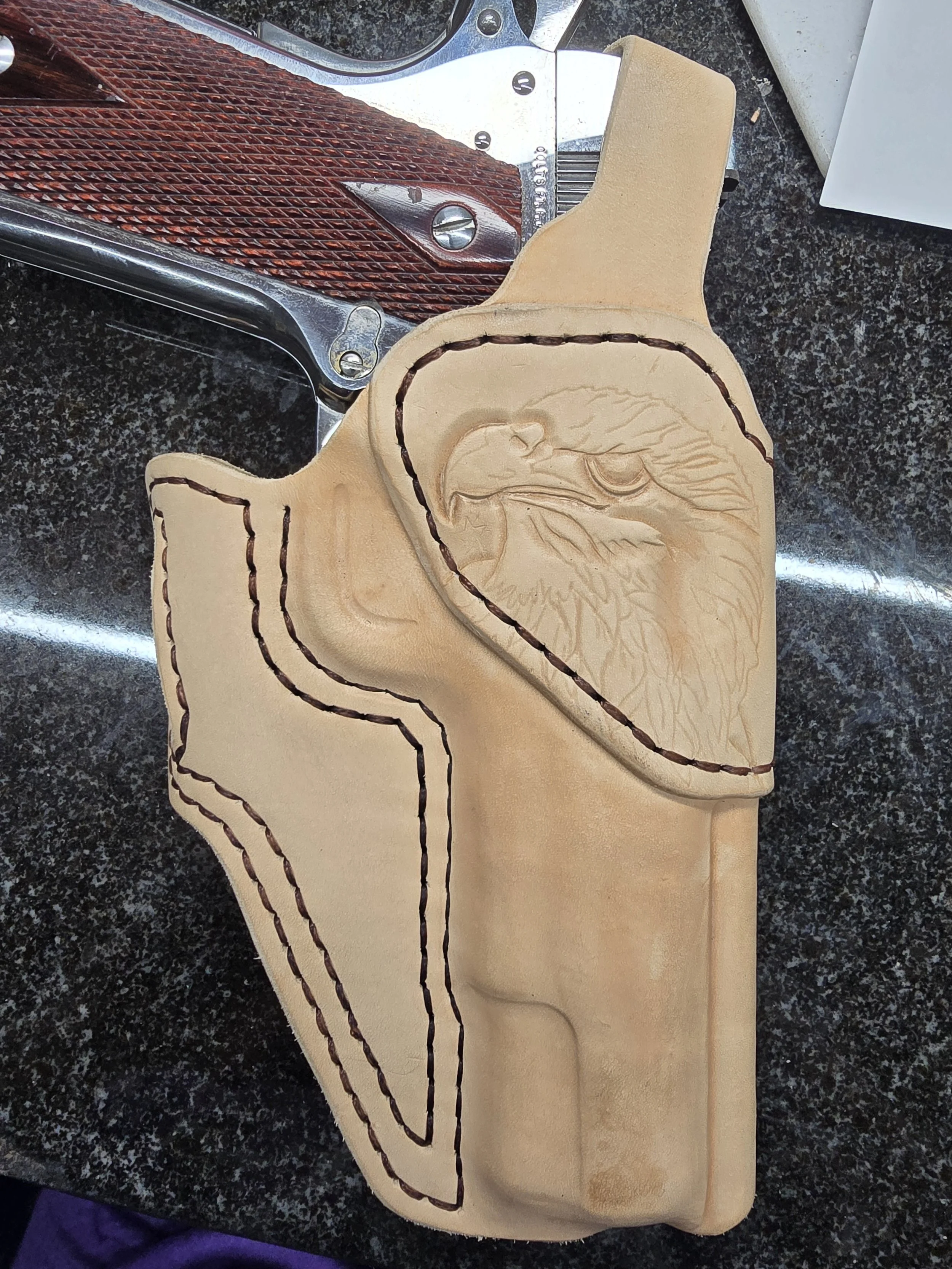 Leather wallet with an engraved eagle design, outlined with dark stitching, placed on a black speckled surface with a metal object and part of a white box nearby.