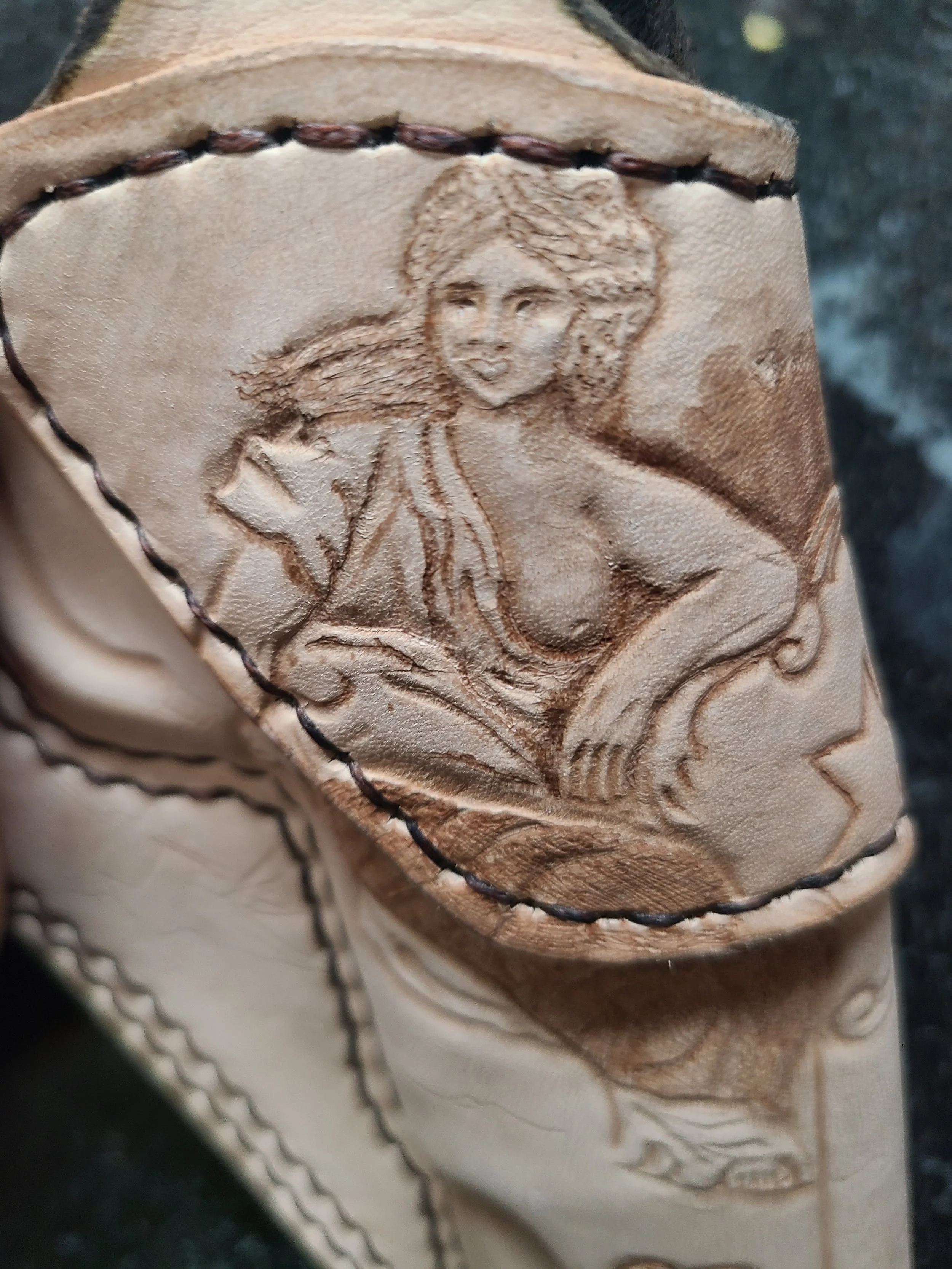 Detailed leather carving of a woman with flowing hair, wearing a low-cut top, sitting with one hand on her hip, in a western-style leather belt with scalloped edges.