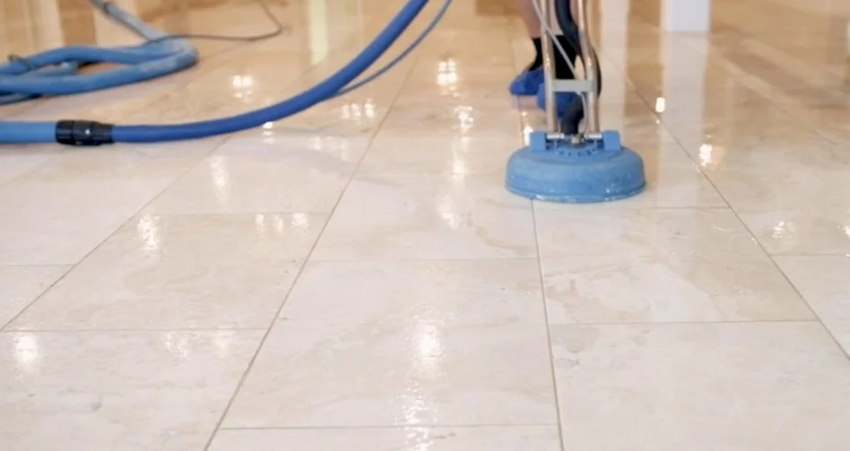 A floor cleaning machine polishes beige marble tiles in a building lobby or hallway.