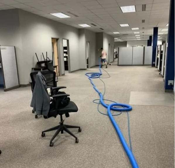 Empty office space with chairs, some stacked, and a person in the background with blue hoses on the carpet.