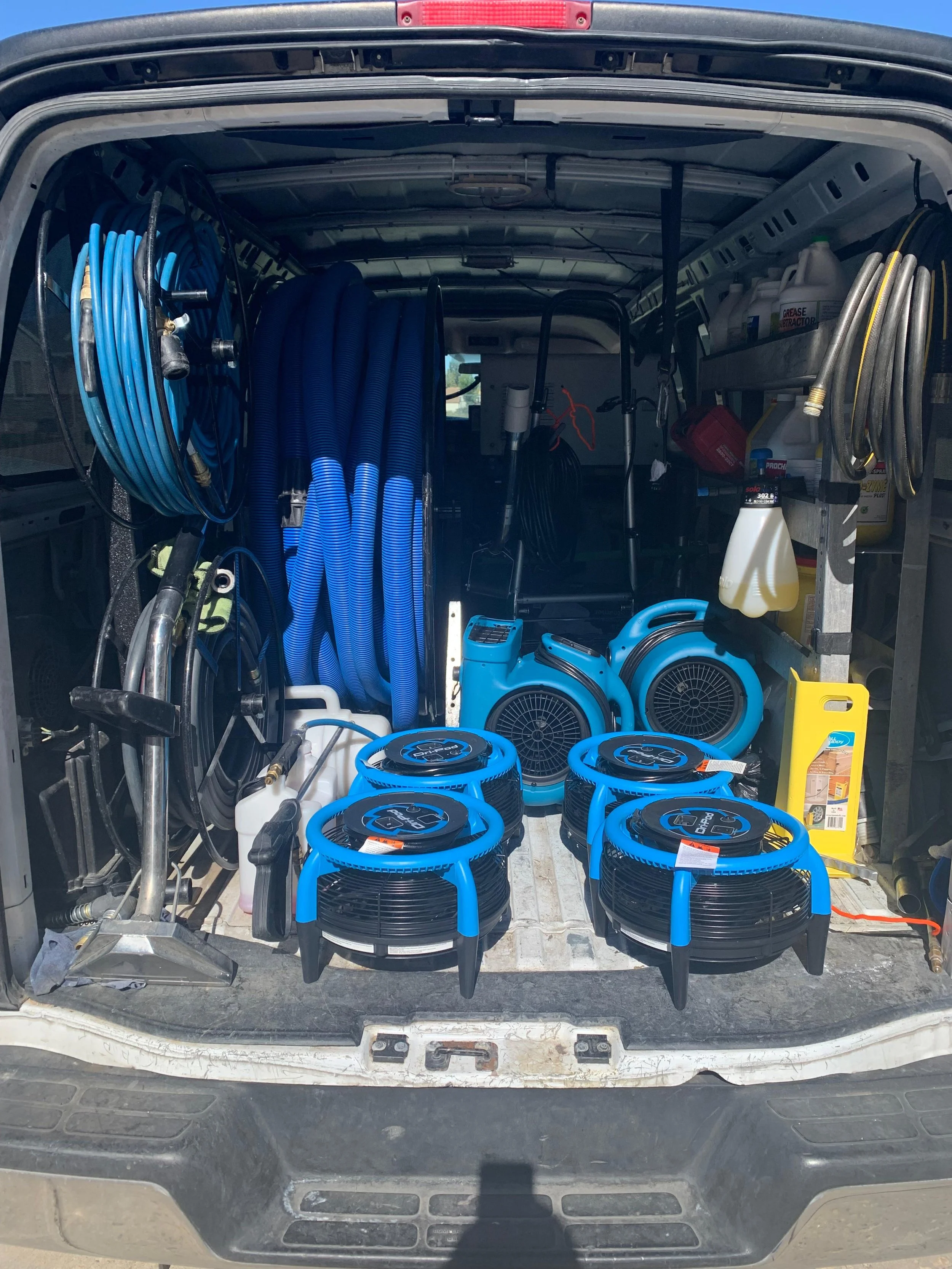 The back of a service van filled with cleaning and industrial equipment, including hoses, fans, and cleaning supplies.