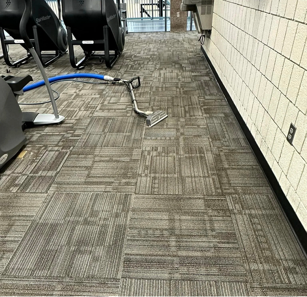 A carpeted floor in an indoor gym or fitness center with cleaning equipment, including a vacuum or floor scrubber, lying on the floor near exercise machines and a brick wall.