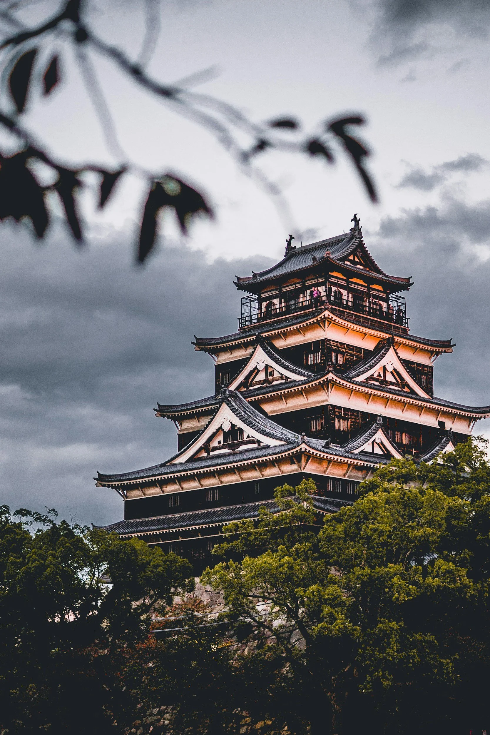 Traditional multi-story Japanese castle with tiered roofs, surrounded by green trees and set against a cloudy sky.