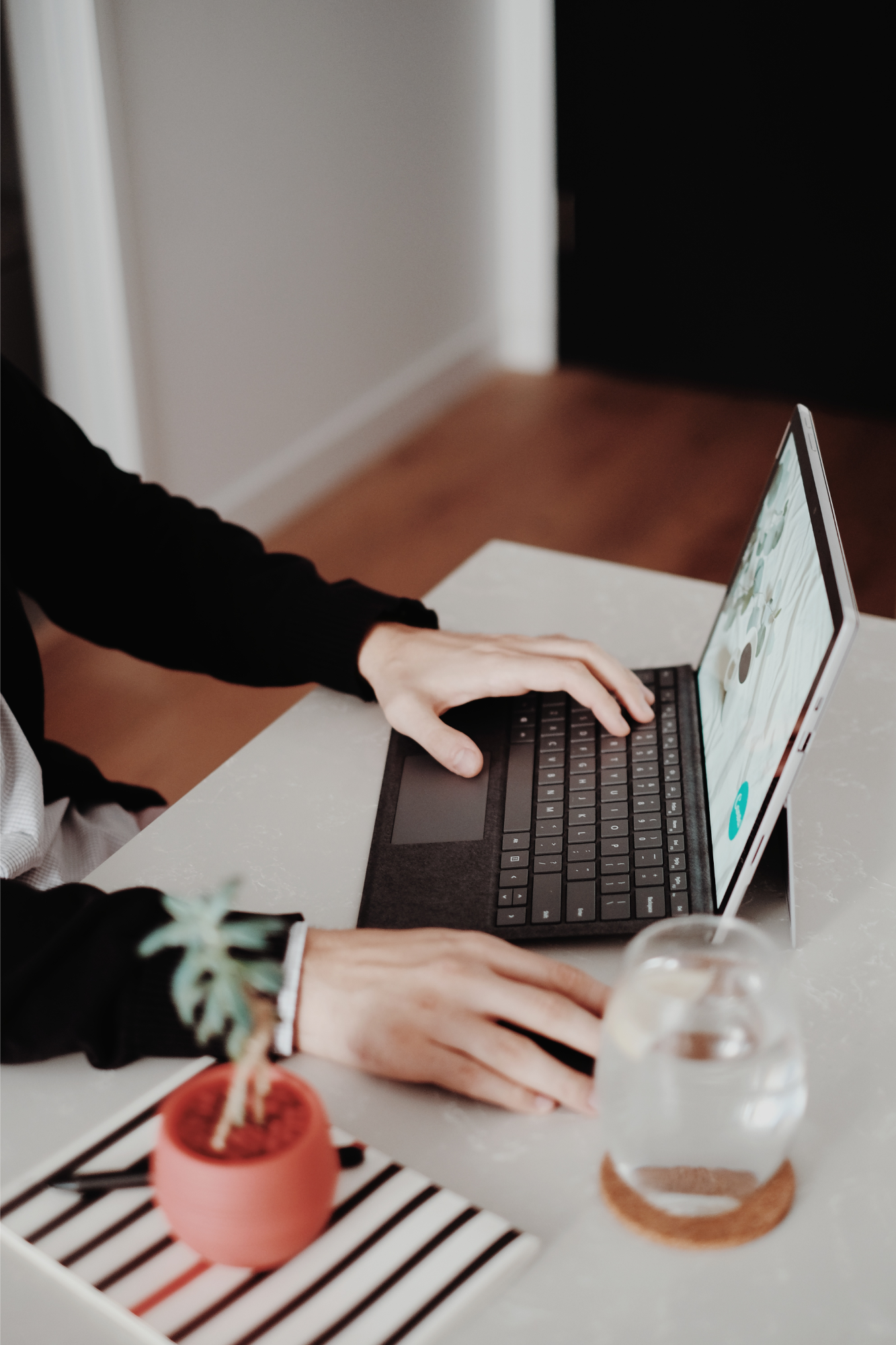 A person working on a laptop doing virtual counseling at a white table, with a small potted plant and a glass of water nearby.