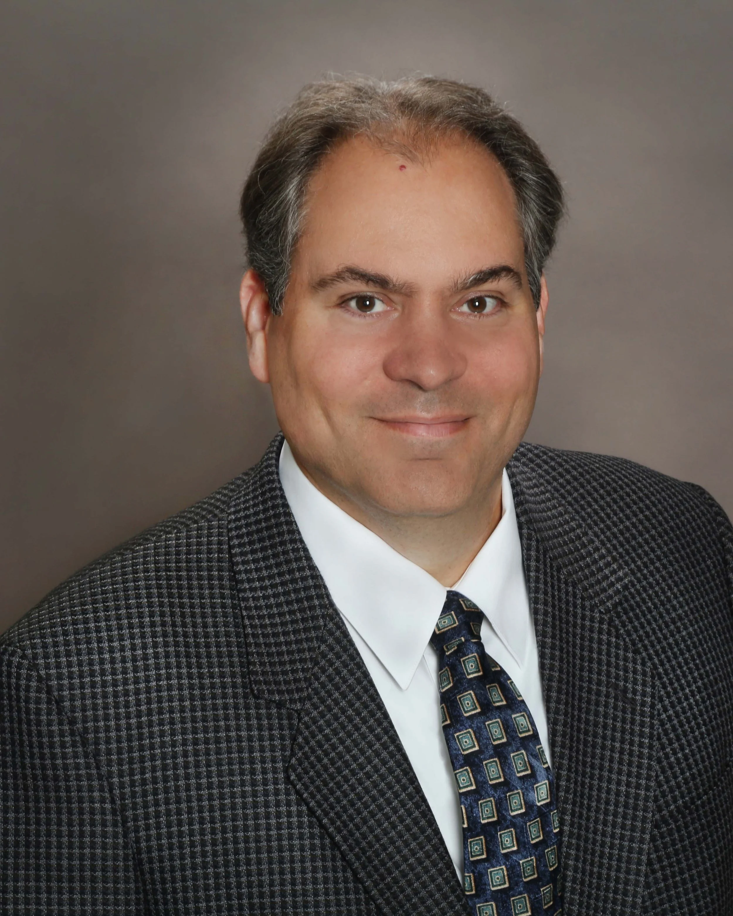 Professional headshot of a man with dark hair, wearing a dark patterned suit, white shirt, and patterned tie, against a neutral background.