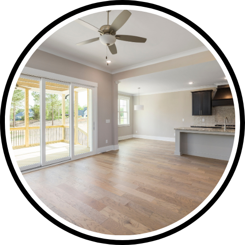 Empty living room with large sliding glass door, ceiling fan, hardwood floors, and an open kitchen with dark cabinets and a white counter.