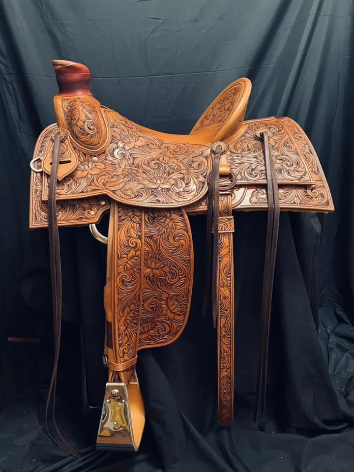 A detailed western saddle with intricate floral carvings, leather straps, and a horn on a black fabric background.
