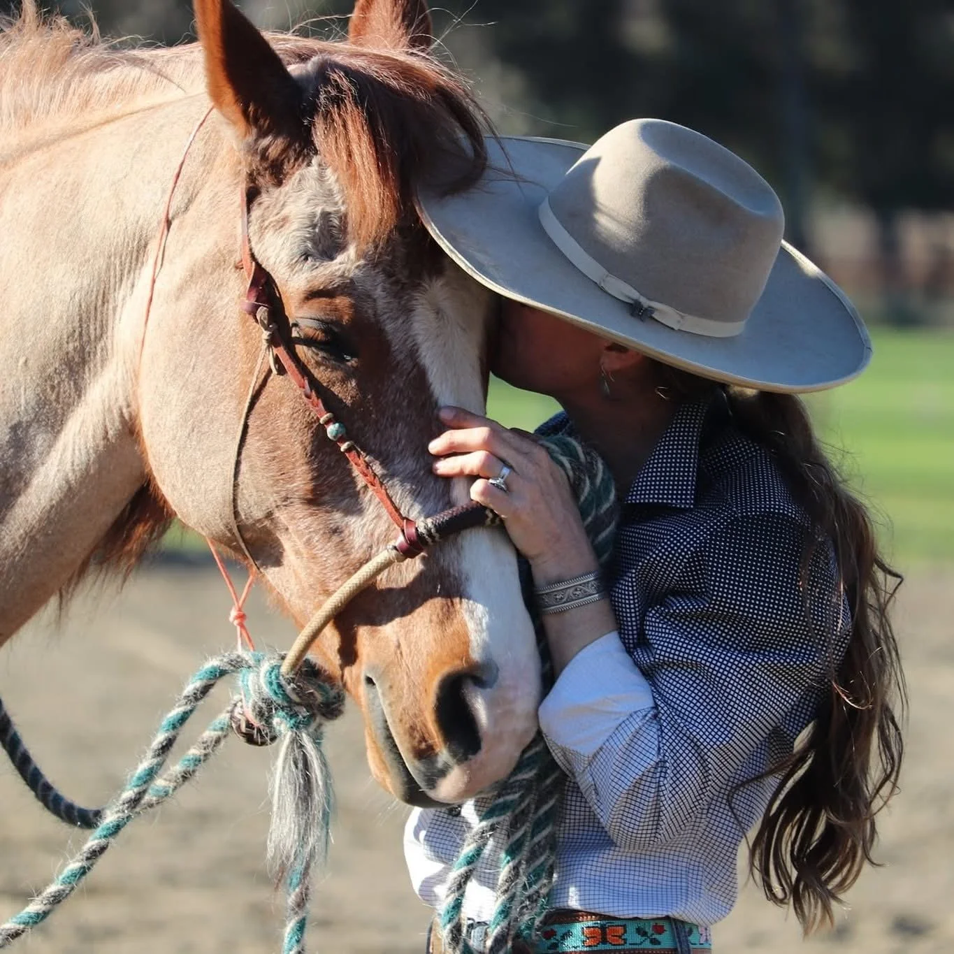 A woman with long hair wearing a large wide-brimmed hat, a patterned shirt, and rings, gently hugging a light tan and white horse with a brown mane in an outdoor setting.