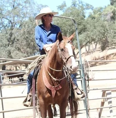 Woman riding a horse while holding a lasso, wearing a cowboy hat and blue shirt, in an outdoor arena with trees.