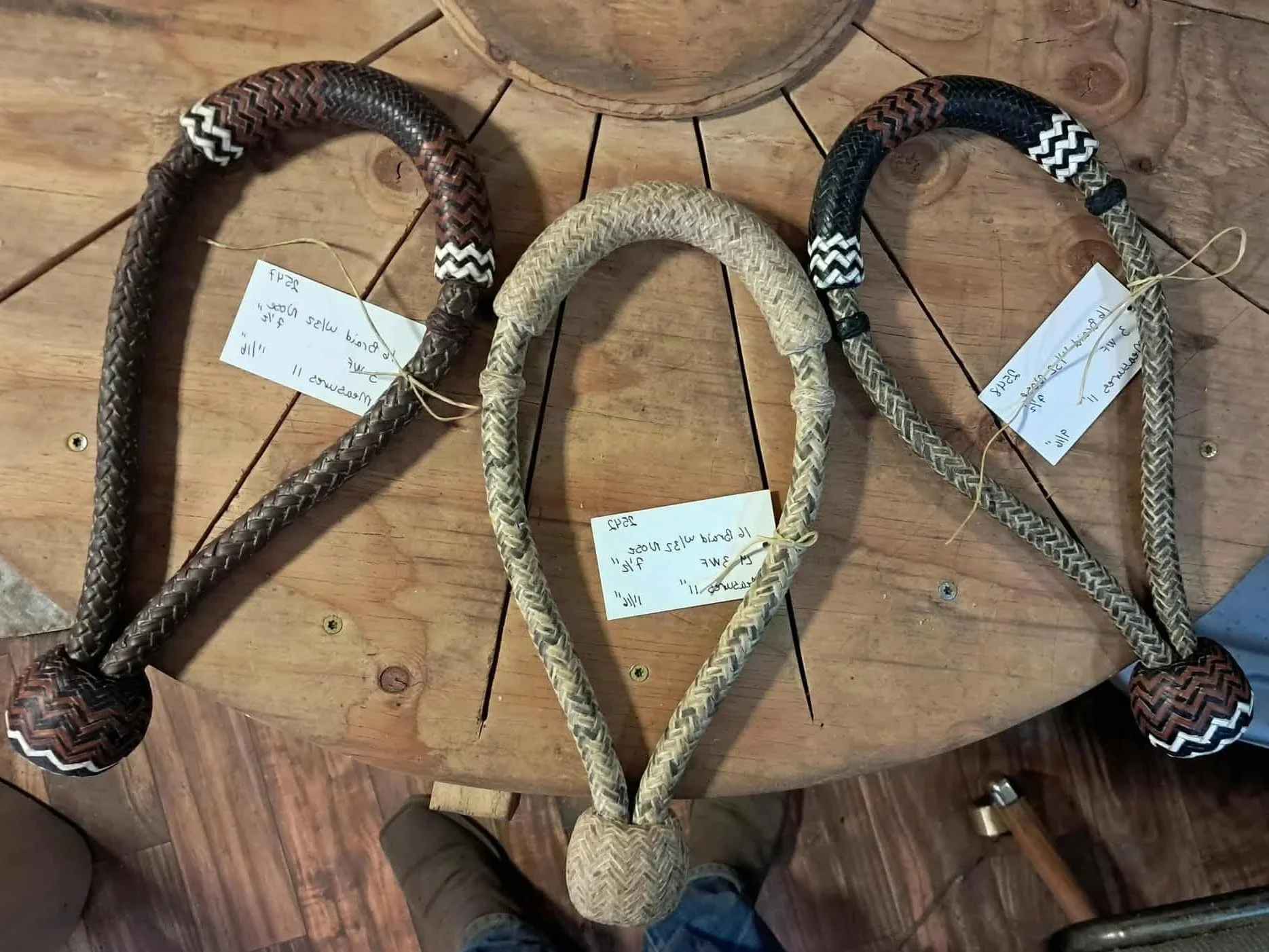 Three woven saddle stirrup straps on a wooden table, each with a handwritten tag.