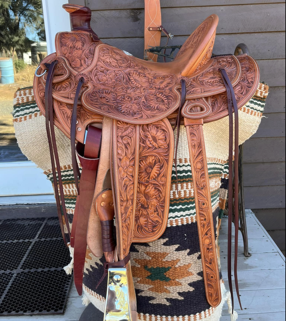 A close-up of a decorated Western-style saddle placed on a woven blanket with geometric patterns, set on a wooden surface outside a building.