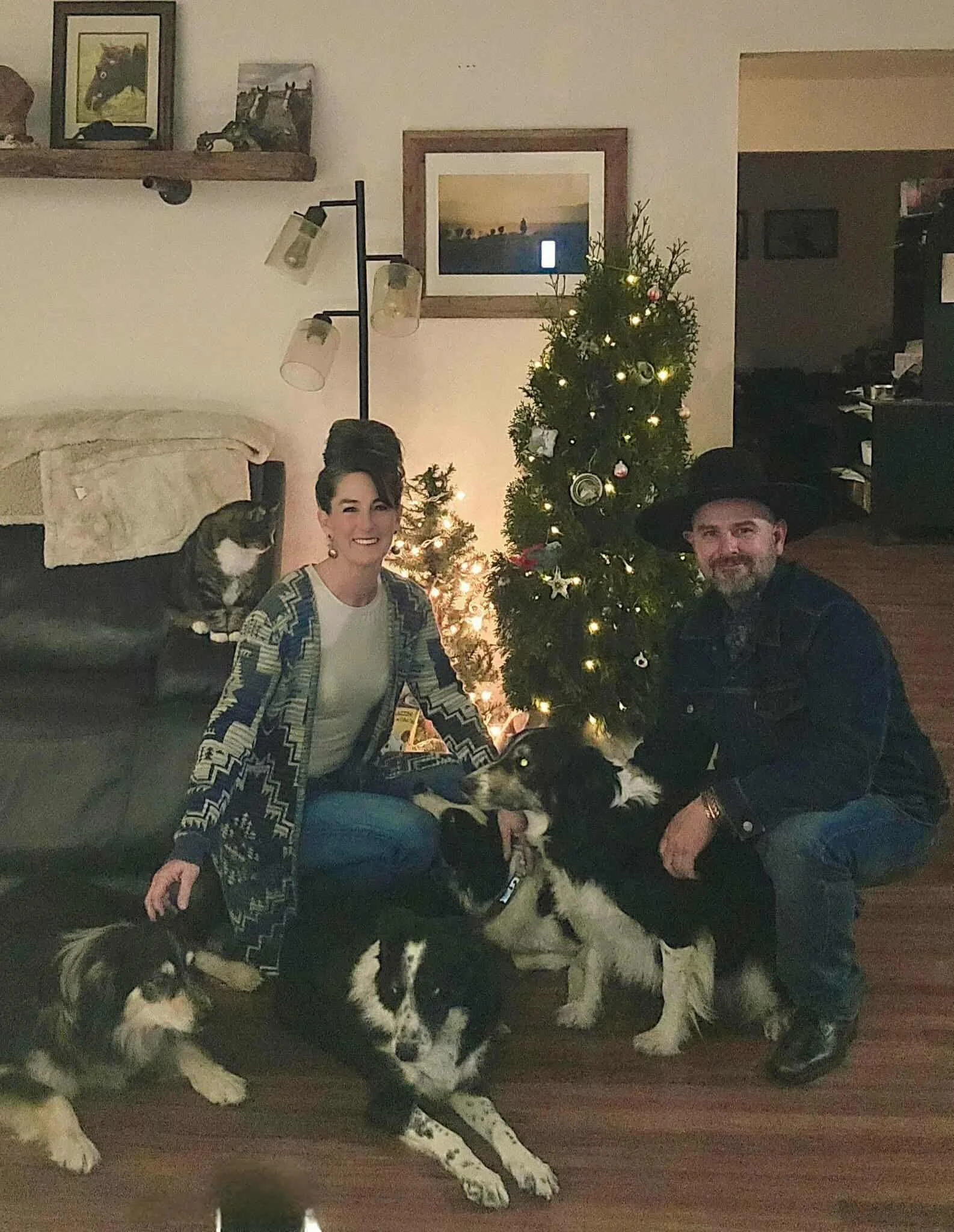 A couple is posing with their dogs in front of a decorated Christmas tree in a living room. The woman is sitting on the floor, smiling, with one dog lying in front of her and another dog sitting beside her. The man is crouching on the right, smiling, with one dog in front of him. There are several other dogs in the scene, and a cat is sitting on a couch on the left. The room has framed pictures and a floor lamp.