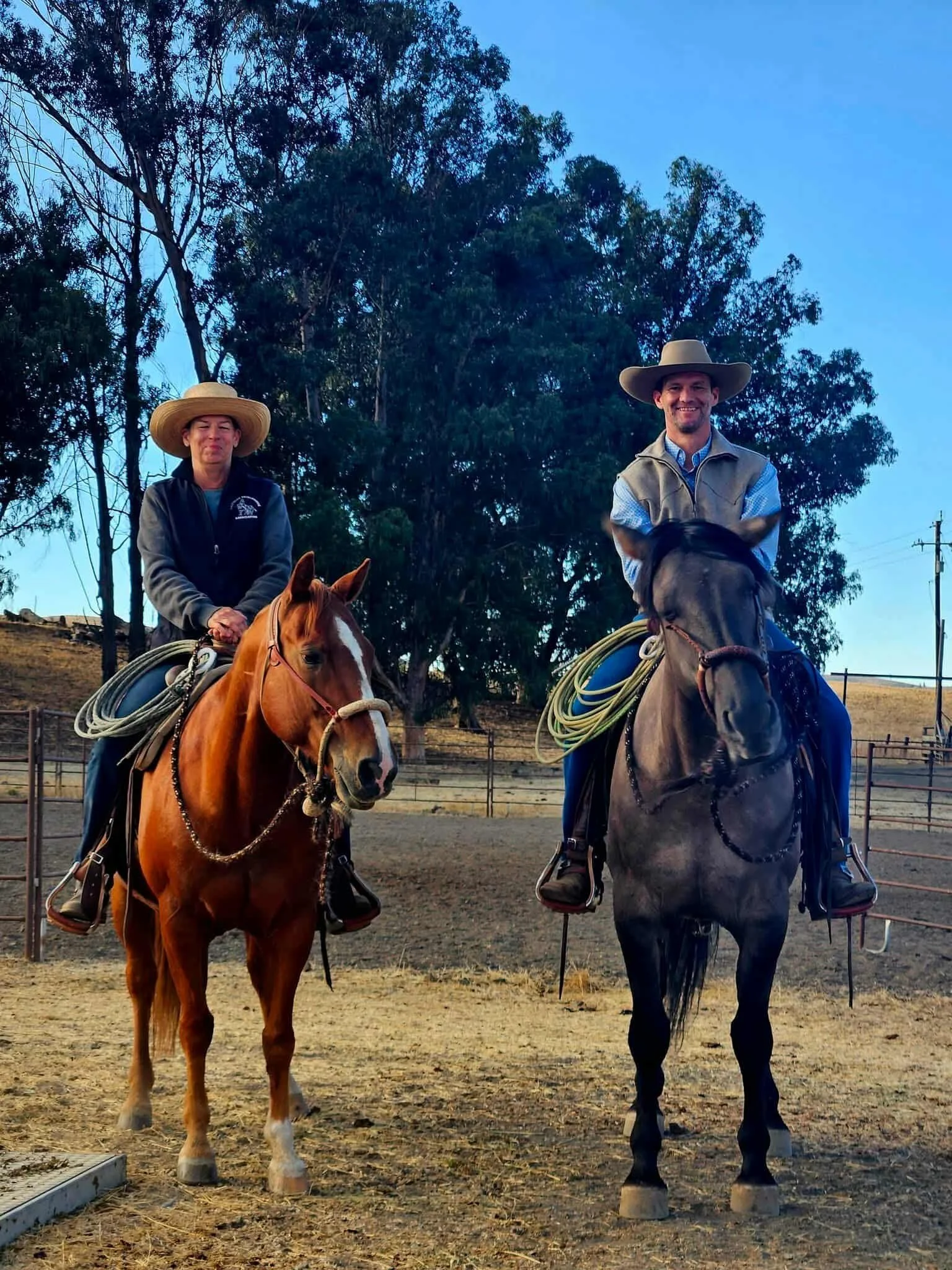 Two people riding horses in an outdoor arena, surrounded by trees and fencing, with clear blue sky overhead.