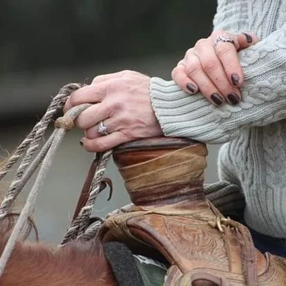 Close-up of a person tightening a leather strap on a saddle, with hands showing a ring and painted nails, wearing a sweater.