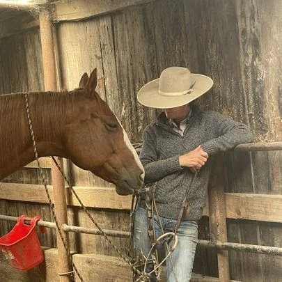 A person wearing a large cowboy hat and gray sweater standing next to a brown horse inside a wooden stable.