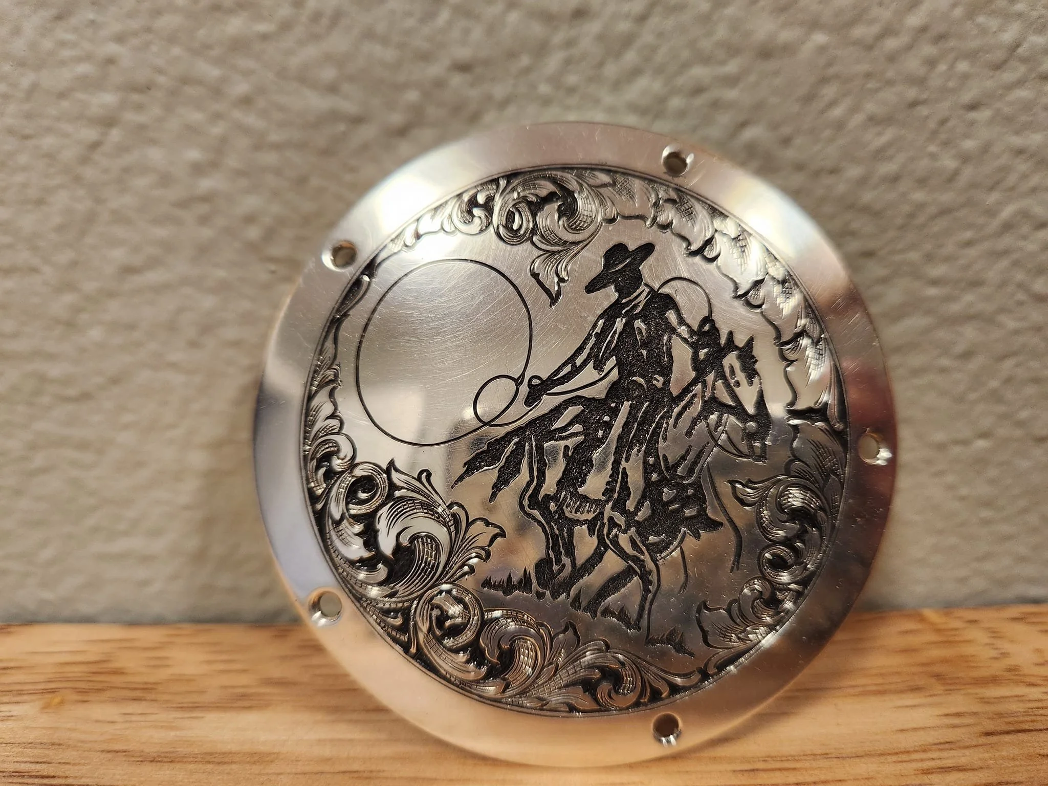 A round, metallic decorative plate featuring a detailed engraved design of a cowboy riding a bucking horse, with swirling patterns and clouds in the background.