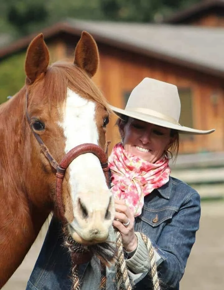 A woman wearing a large white hat, pink floral scarf, and denim jacket smiling and holding a brown and white horse with a halter at a farm.