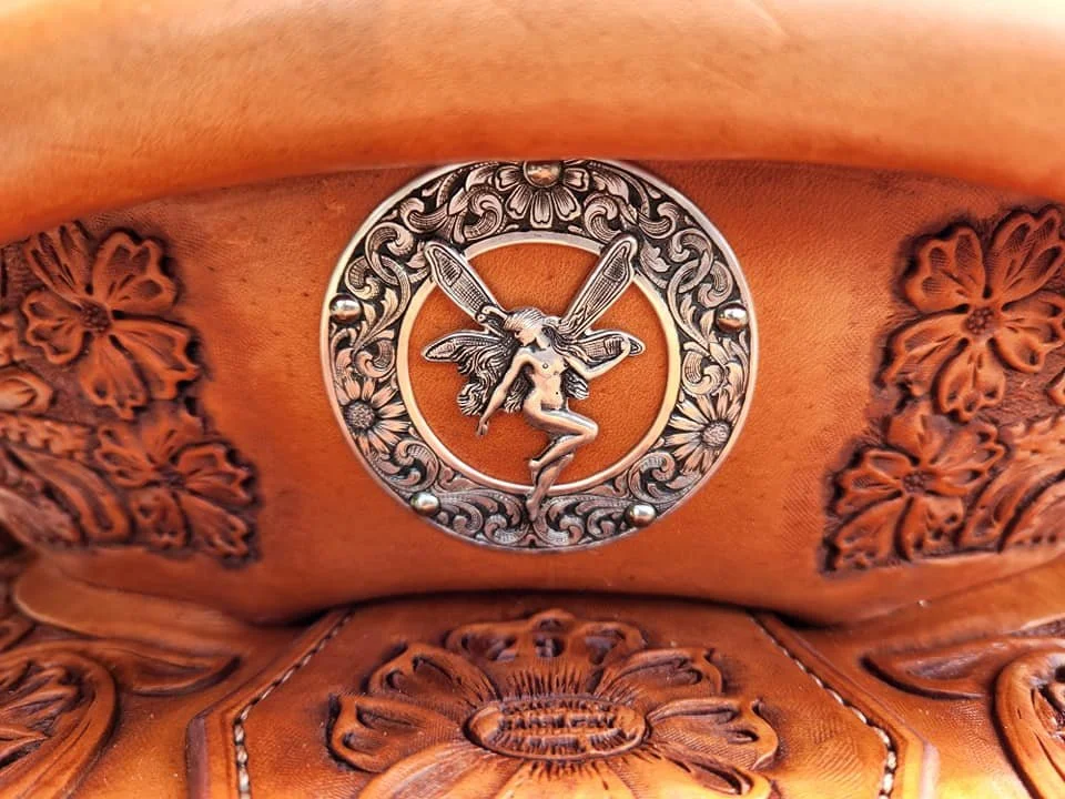 Close-up of a decorative silver buckle featuring a fairy with wings, surrounded by floral and scroll engravings, attached to brown leather with floral tooled designs.