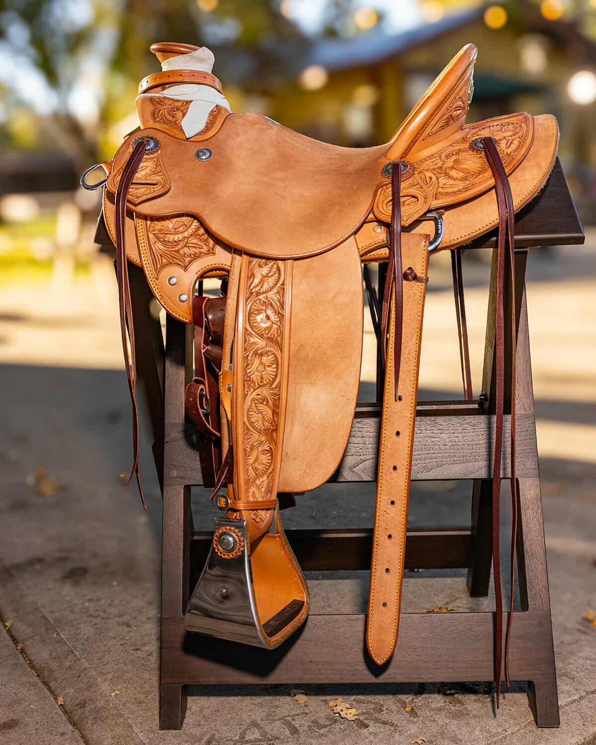 Leather western saddle with intricate carving details placed on a wooden saddle stand outdoors.