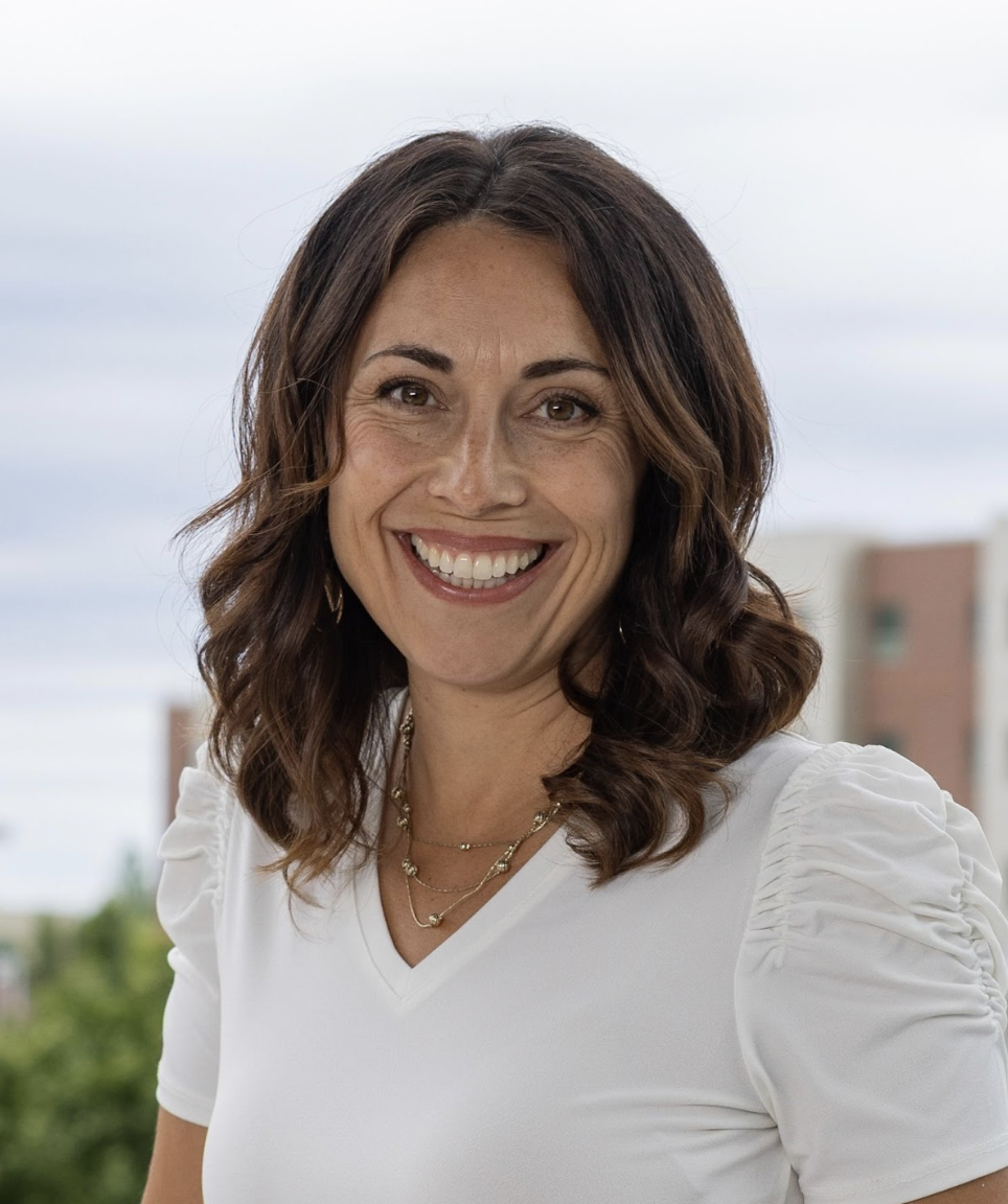 A smiling woman with shoulder-length brown hair, wearing a white top with puffed sleeves and layered necklaces, standing outdoors with a blurred background of trees and buildings.
