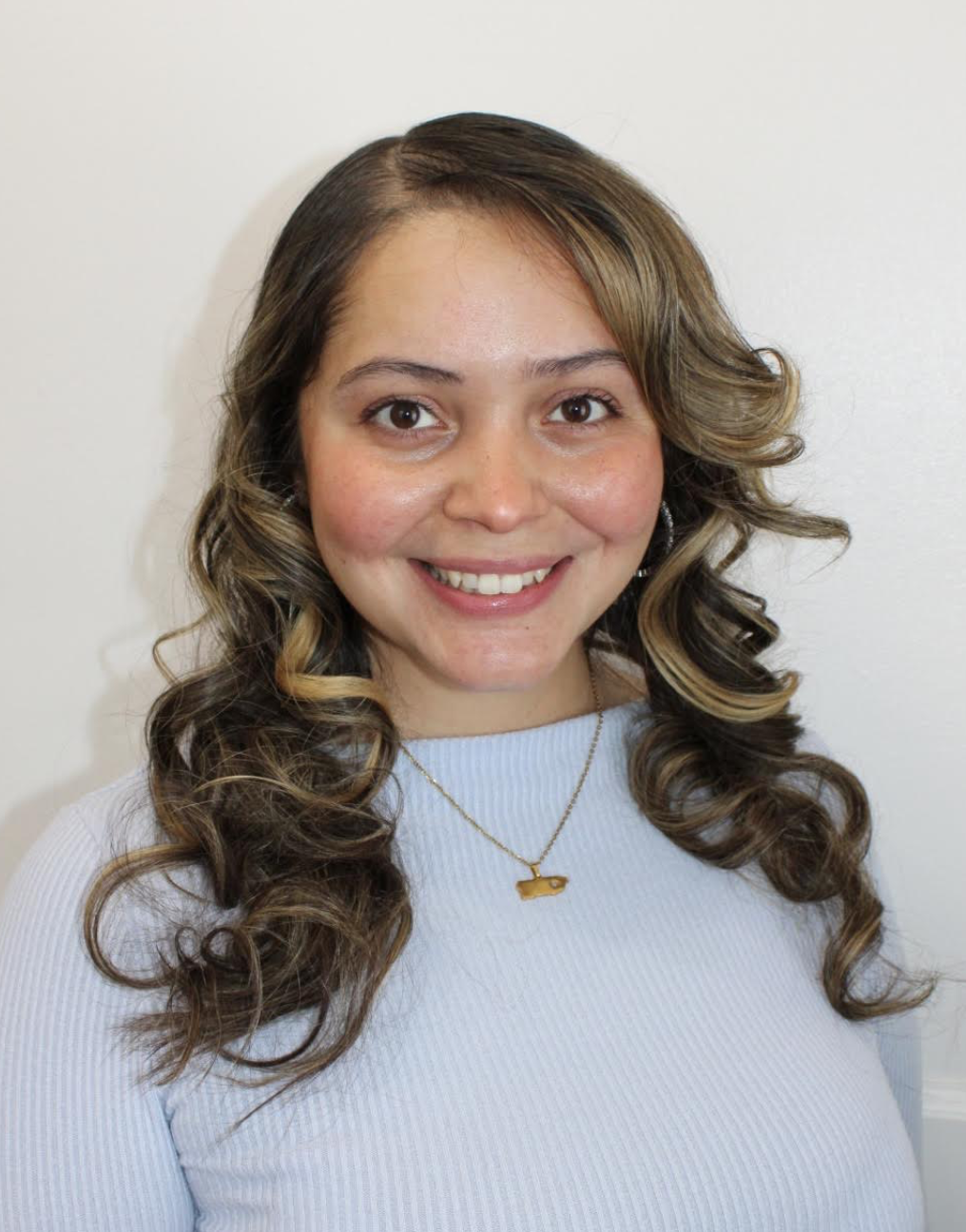 A smiling woman with brown, curled hair wearing a light blue top and a gold necklace.