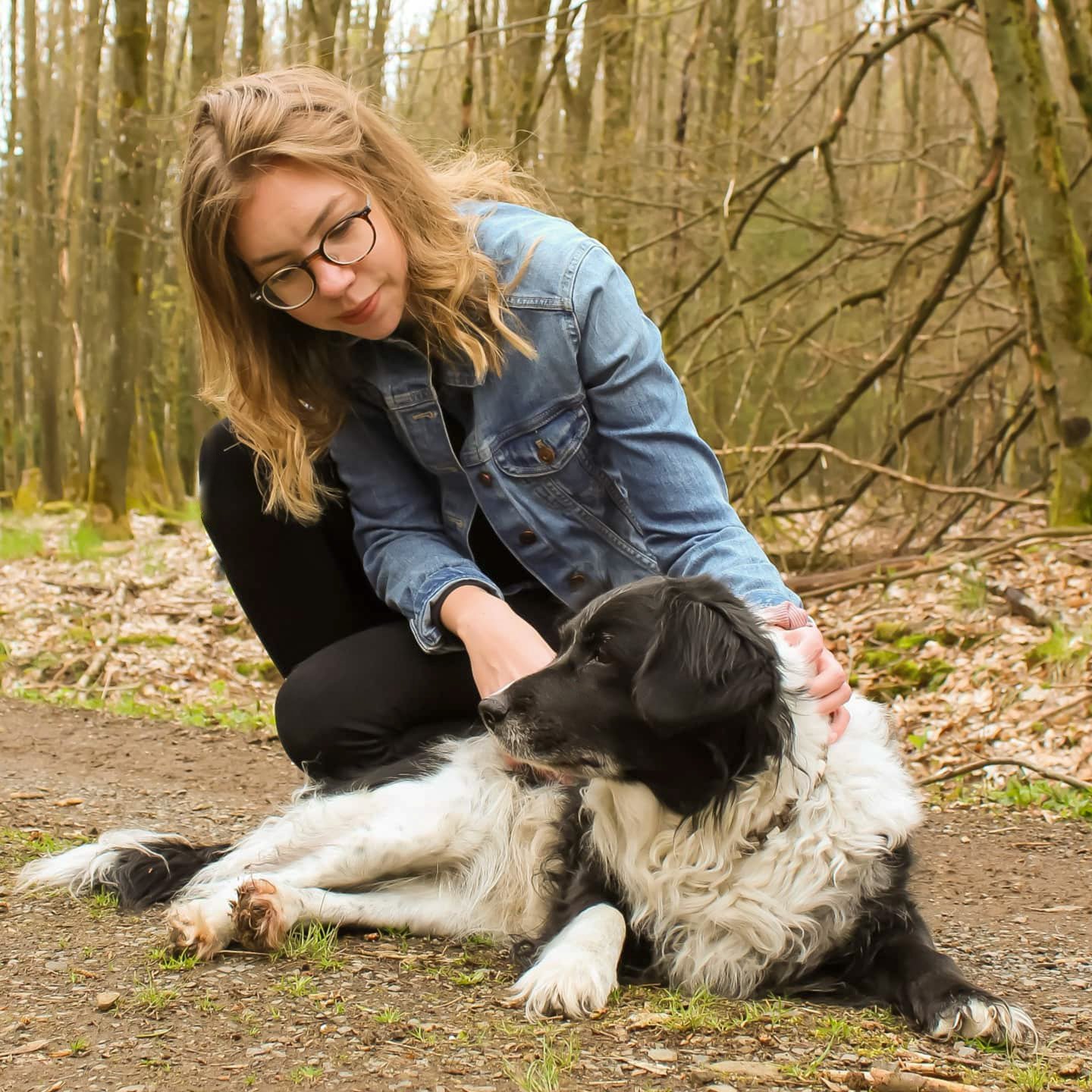 Eine junge Frau mit Brille und Hellbraunen Haaren, die in einem Wald einen Hund streichelt, der auf dem Boden liegt.
