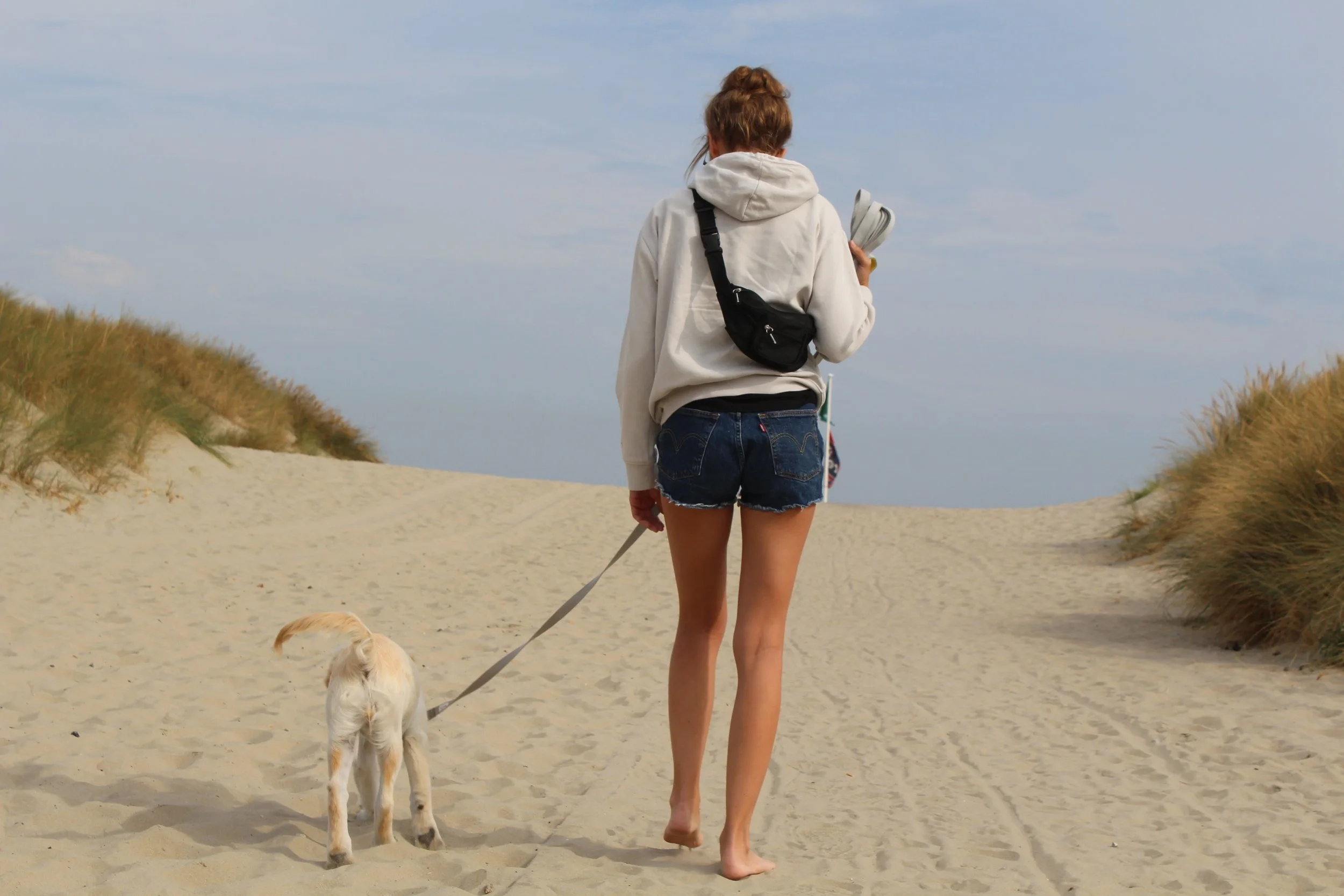 Frau geht barfuß auf Sanddüne mit Hund an Leine entlang am Strand.