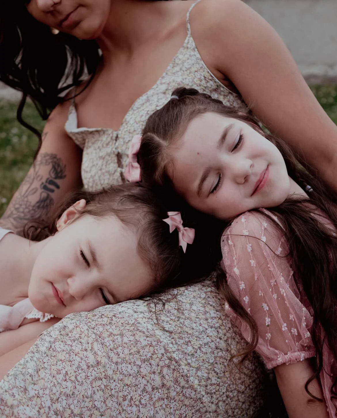 A woman and two young girls are lying down outdoors with their heads close together, all with eyes closed and smiling softly. The woman has dark hair and tattoos on her arm, wearing a light-colored, patterned sleeveless top. The girl in the middle ha