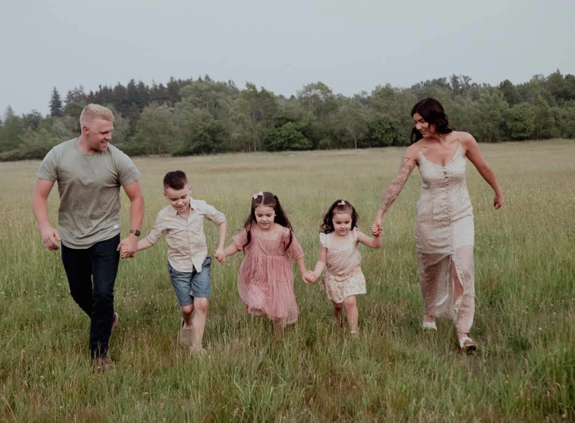 A family of five, including a mother, father, and three young girls, holding hands and walking through a grassy field with trees in the background.
