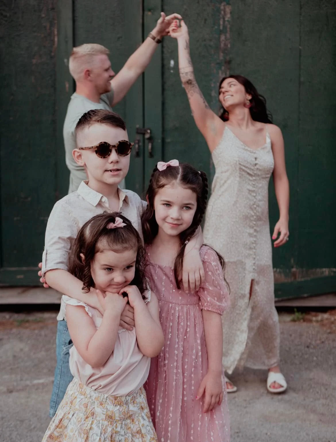 A family photo with four children in the foreground, including a girl in a pink dress and a boy with sunglasses, and two adults dancing in the background in front of a dark green door.