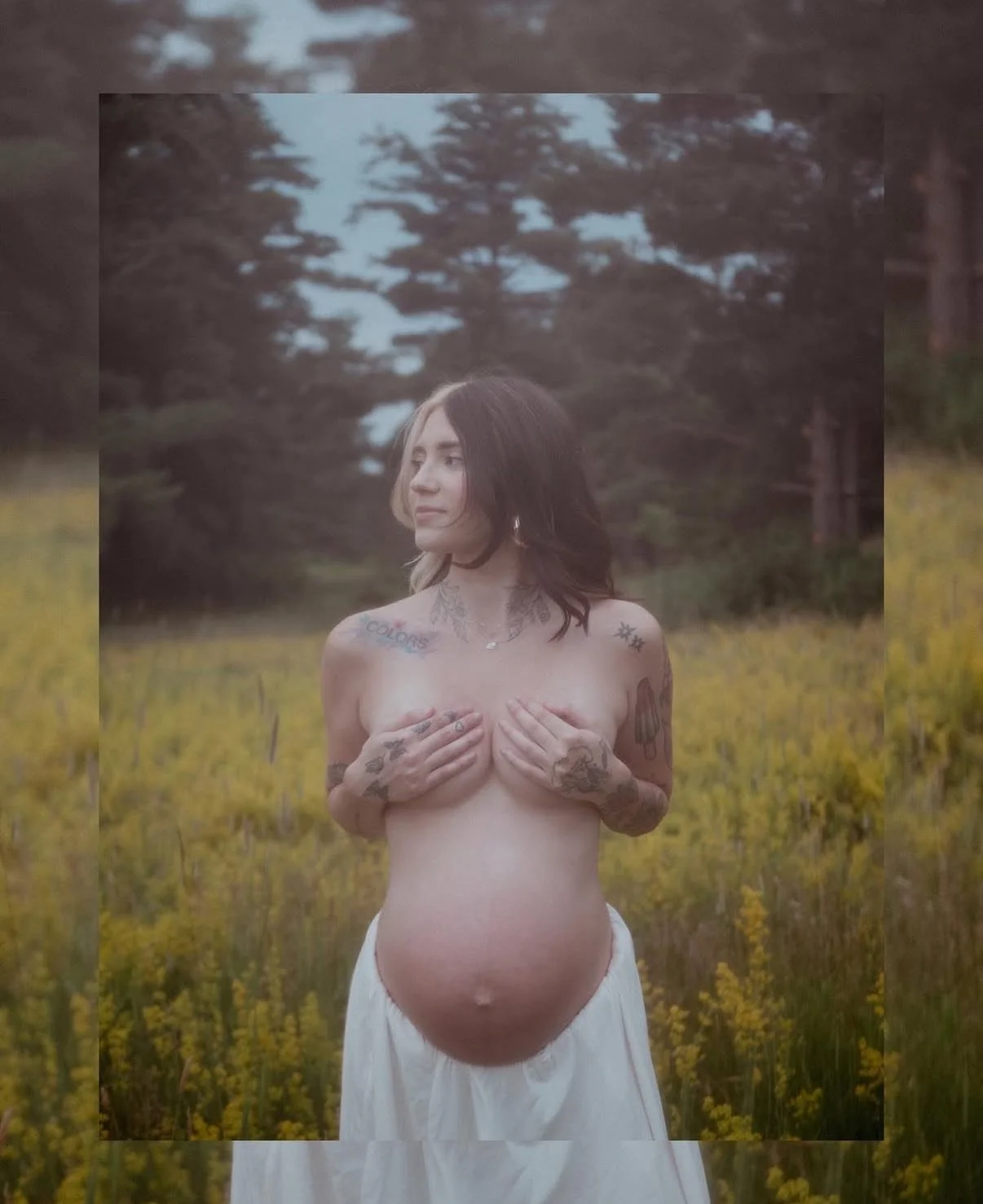 A pregnant woman standing outdoors in a field of yellow flowers, holding her breasts with her hands, with trees in the background.