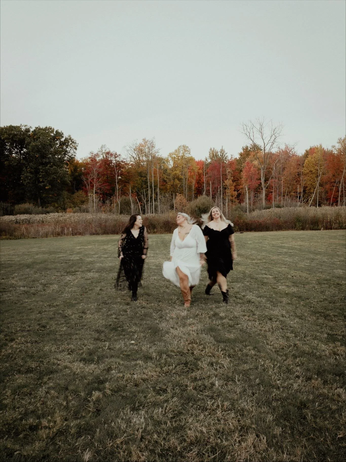 Three women walking and smiling across a grassy field during autumn, with trees in fall colors in the background.