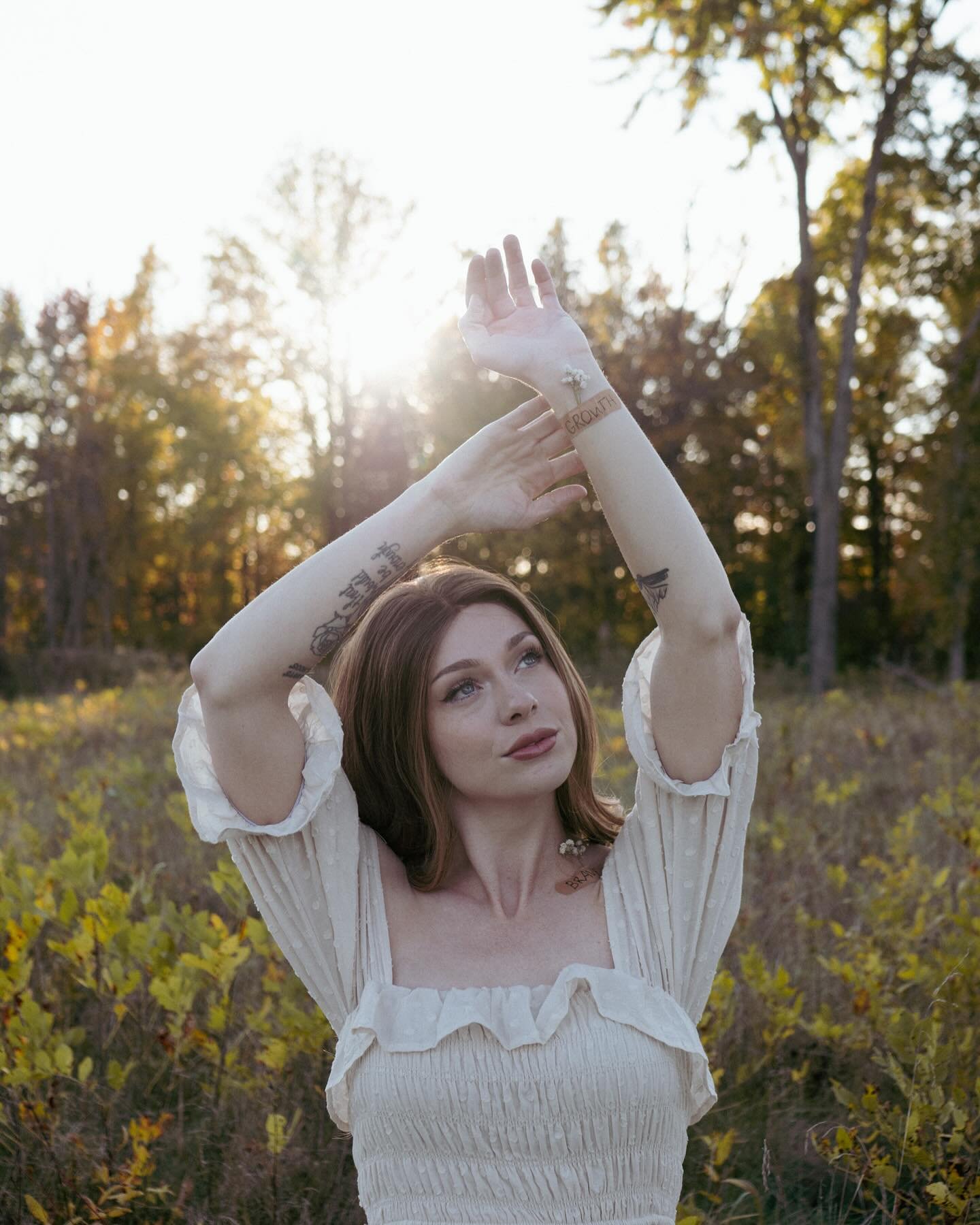 A woman standing outdoors in a field with trees in the background, holding her arms up towards the sunlight during sunset.