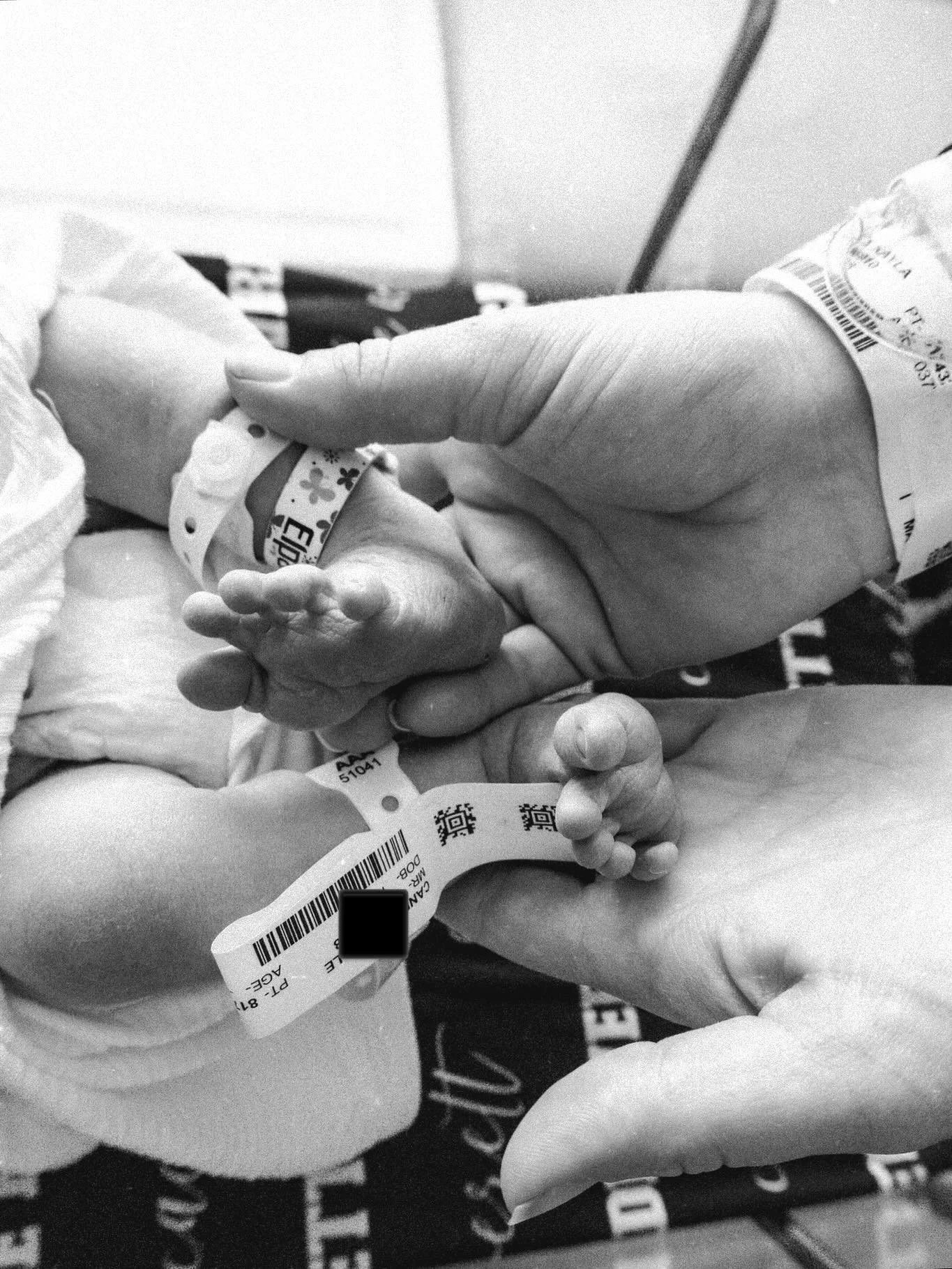 A baby's hand holding an adult's finger, with hospital wristbands visible on both.