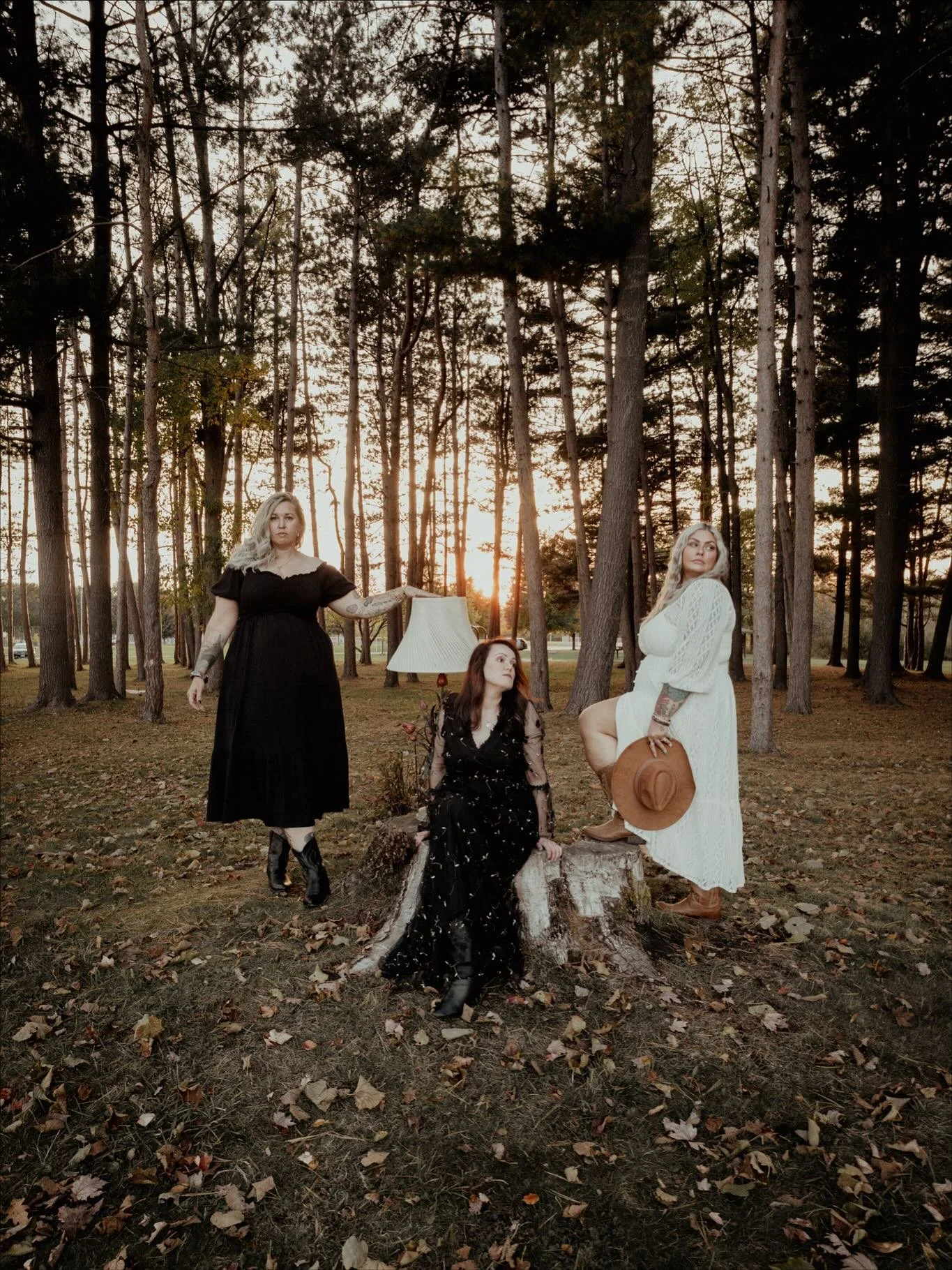Three women in dresses pose outdoors in a wooded area at sunset. One woman in a black dress stands on the left, one woman in a black dress sits on a tree stump in the middle, and one woman in a white dress stands on the right holding a hat.