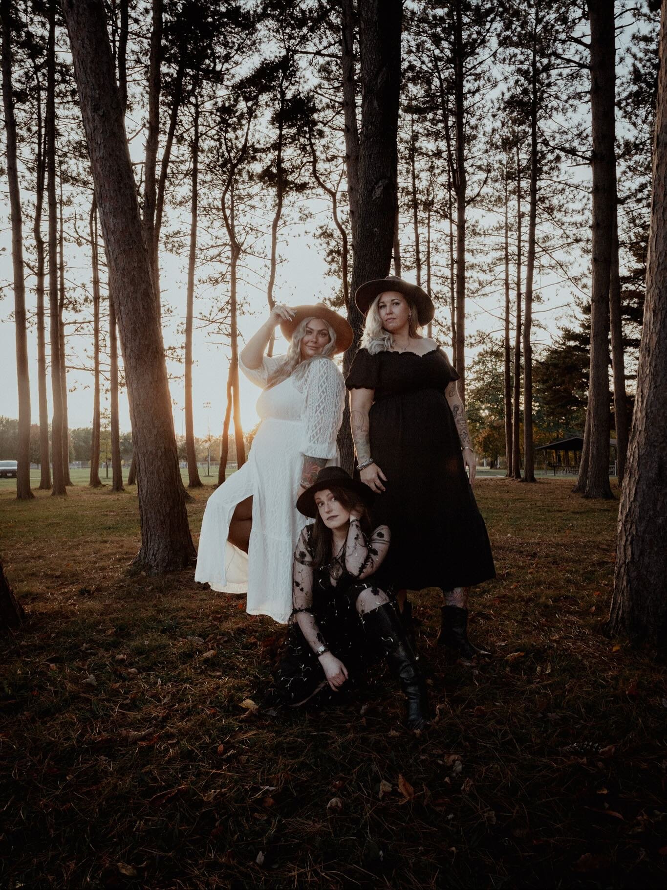 Three women in vintage-style dresses and wide-brimmed hats posing in a forest during sunset.