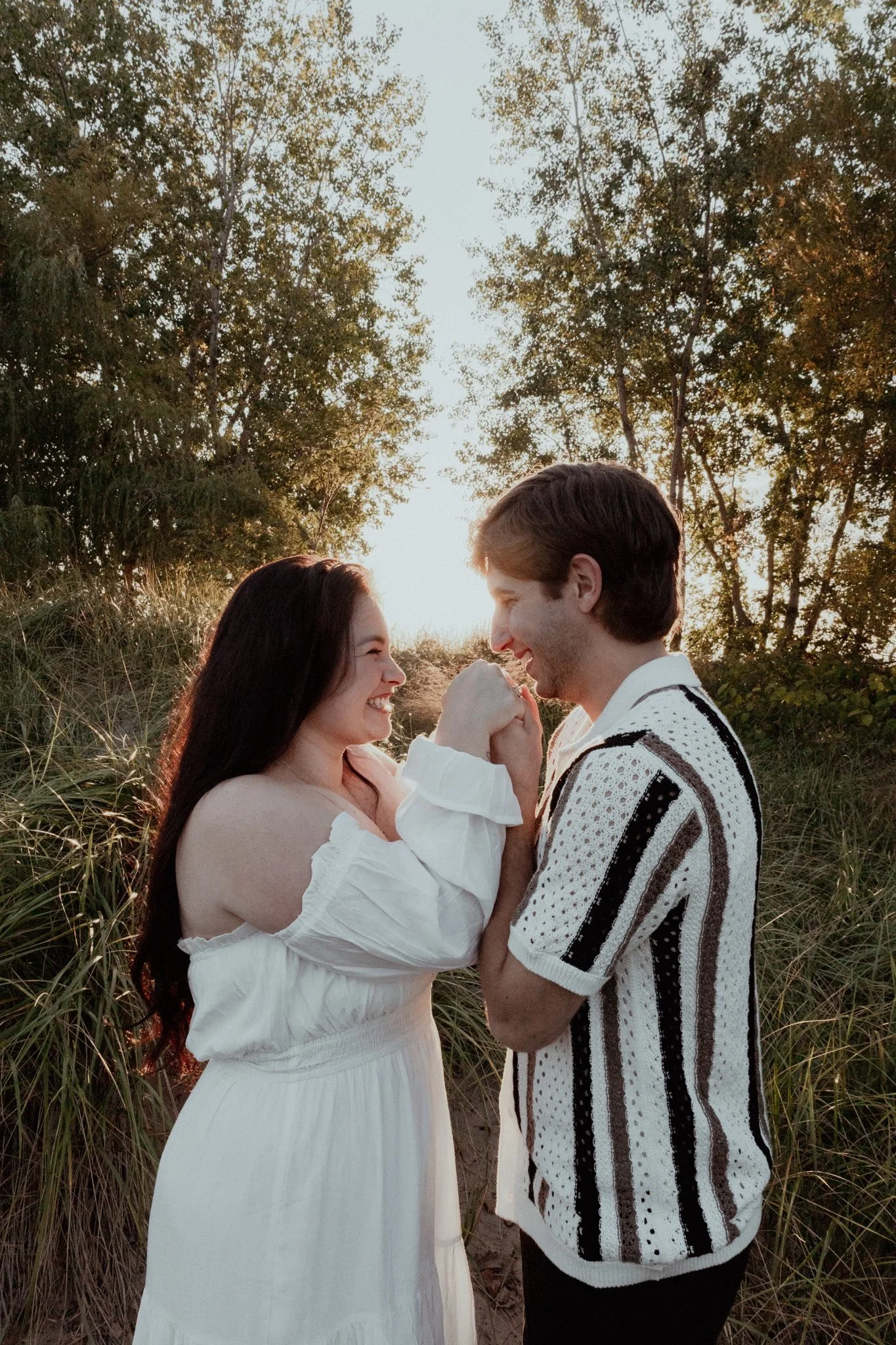 A couple smiling and holding hands in a natural outdoor setting during sunset.