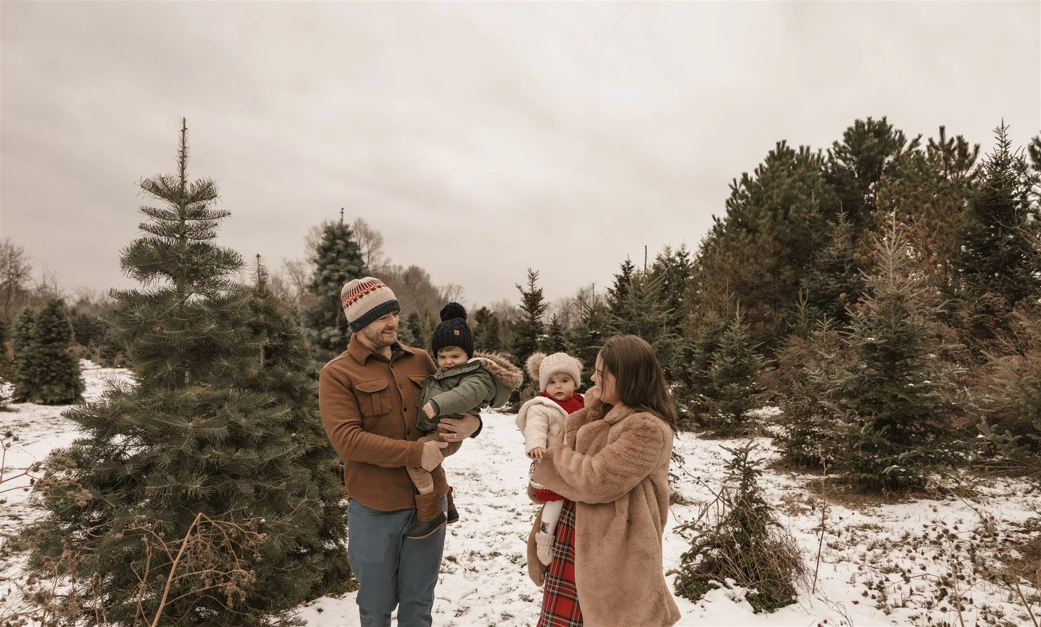Family of four outdoors in a snowy Christmas tree farm, with pine trees in the background, wearing winter clothing, smiling and enjoying the day.