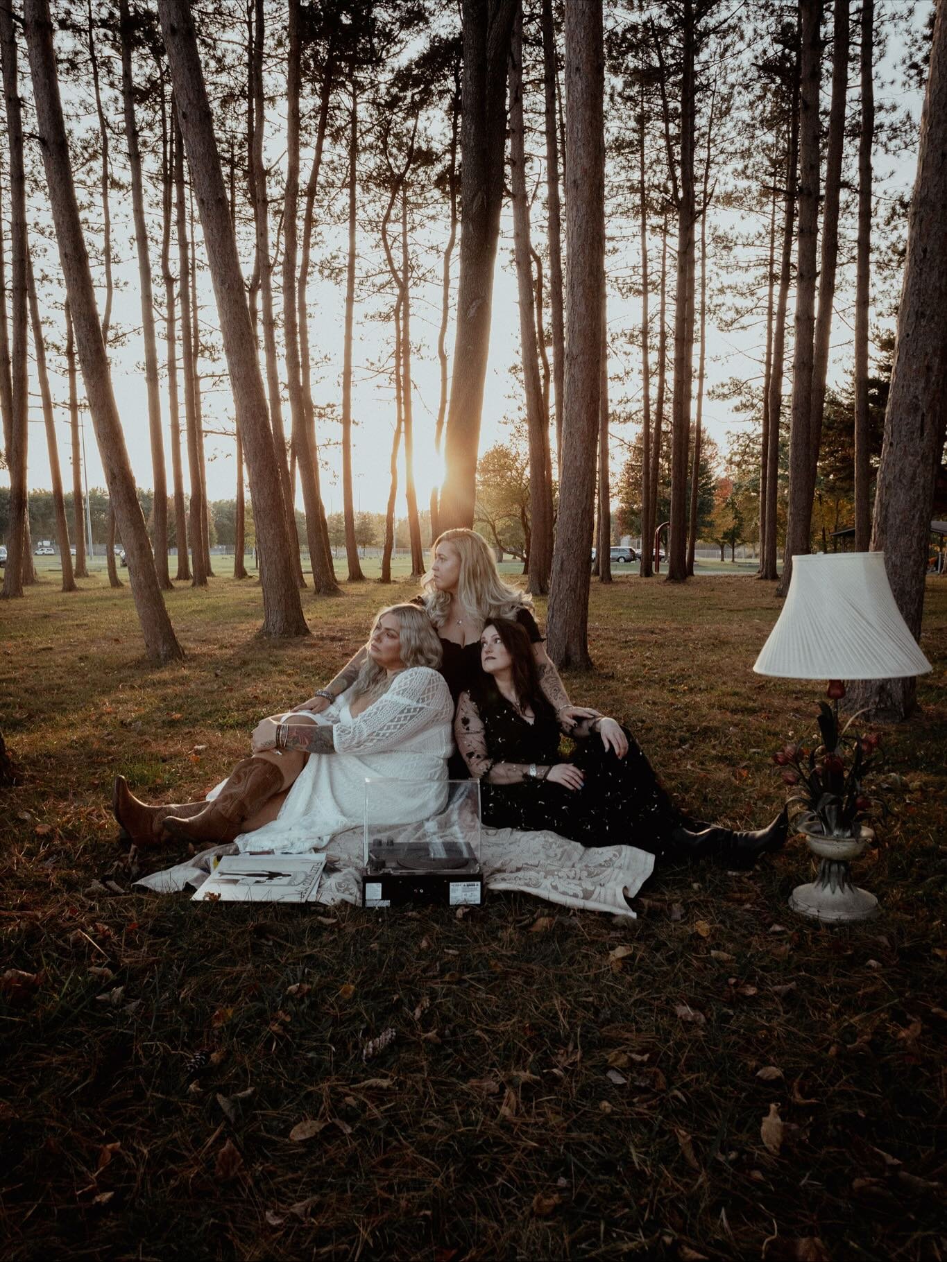 Three women sitting on a blanket in a wooded park during sunset, with a record player and a lamp nearby.