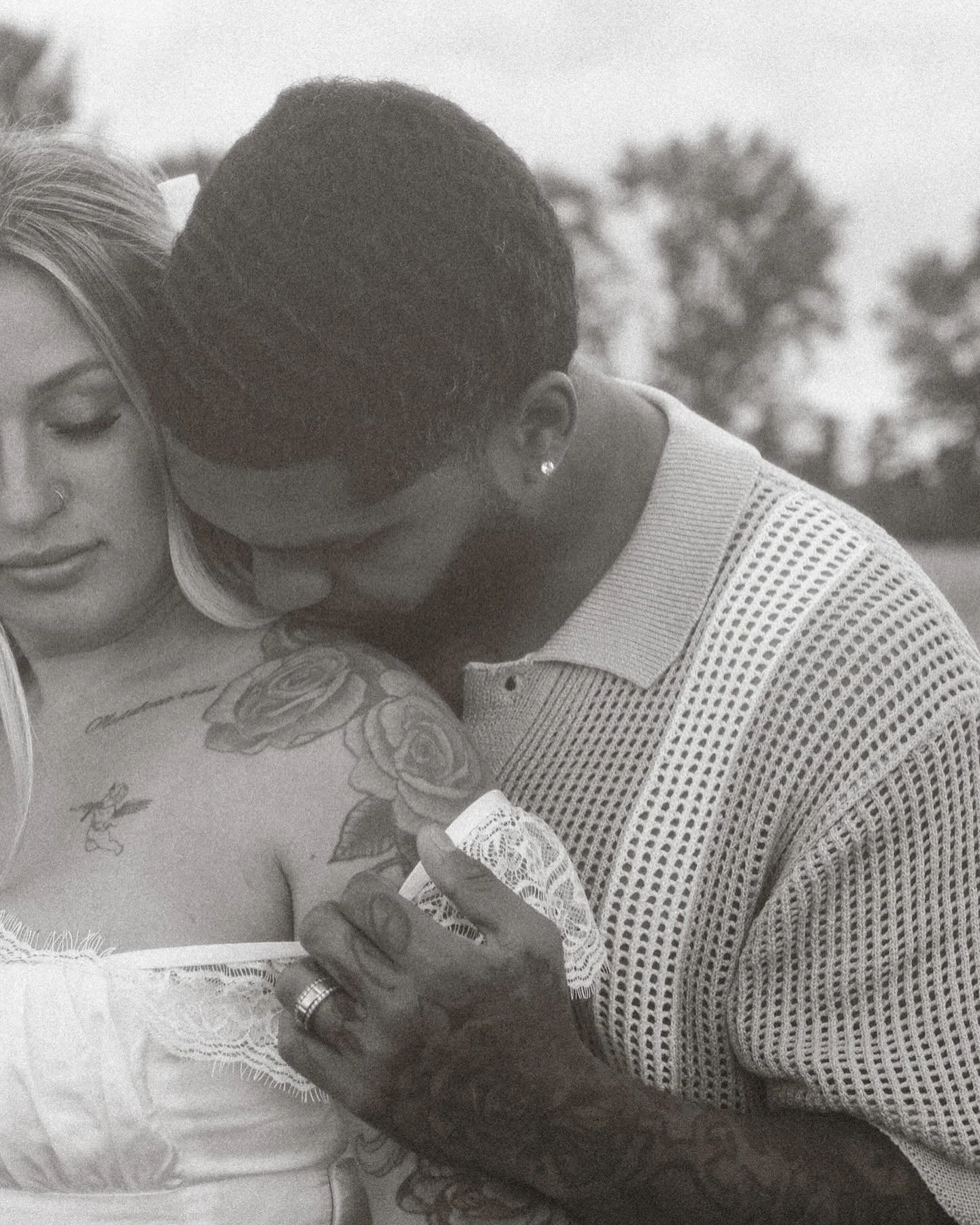 A couple embraces outdoors, with his hand on her shoulder, a man with tattoos and earrings, and a woman with blonde hair and tattoos on her chest.