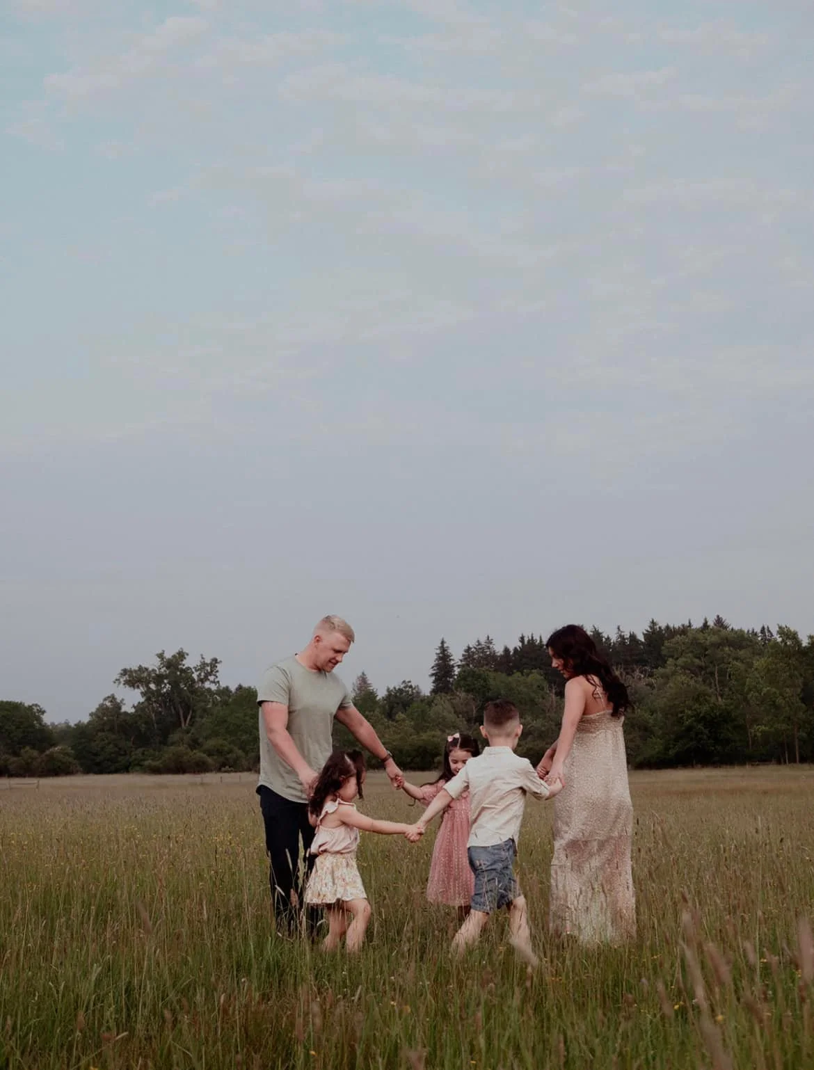 A family of five holding hands in a circle in a grassy field with trees in the background.