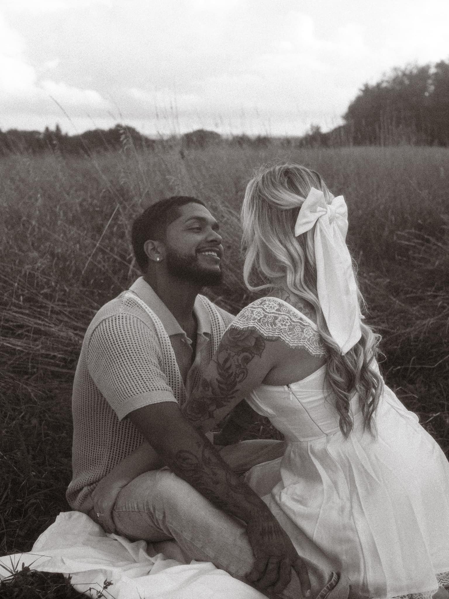 A black and white photo of a couple sitting outdoors in a field, smiling and looking at each other. The woman has long wavy hair with a large bow and tattoos on her arm, wearing a white dress. The man has a beard, short hair, and tattoos on his arm, 