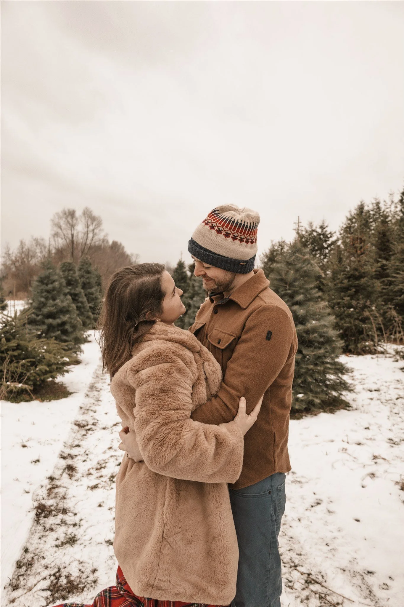 A couple stands close together in a snowy outdoor Christmas tree farm, gazing into each other's eyes, dressed warmly in winter coats and hats.