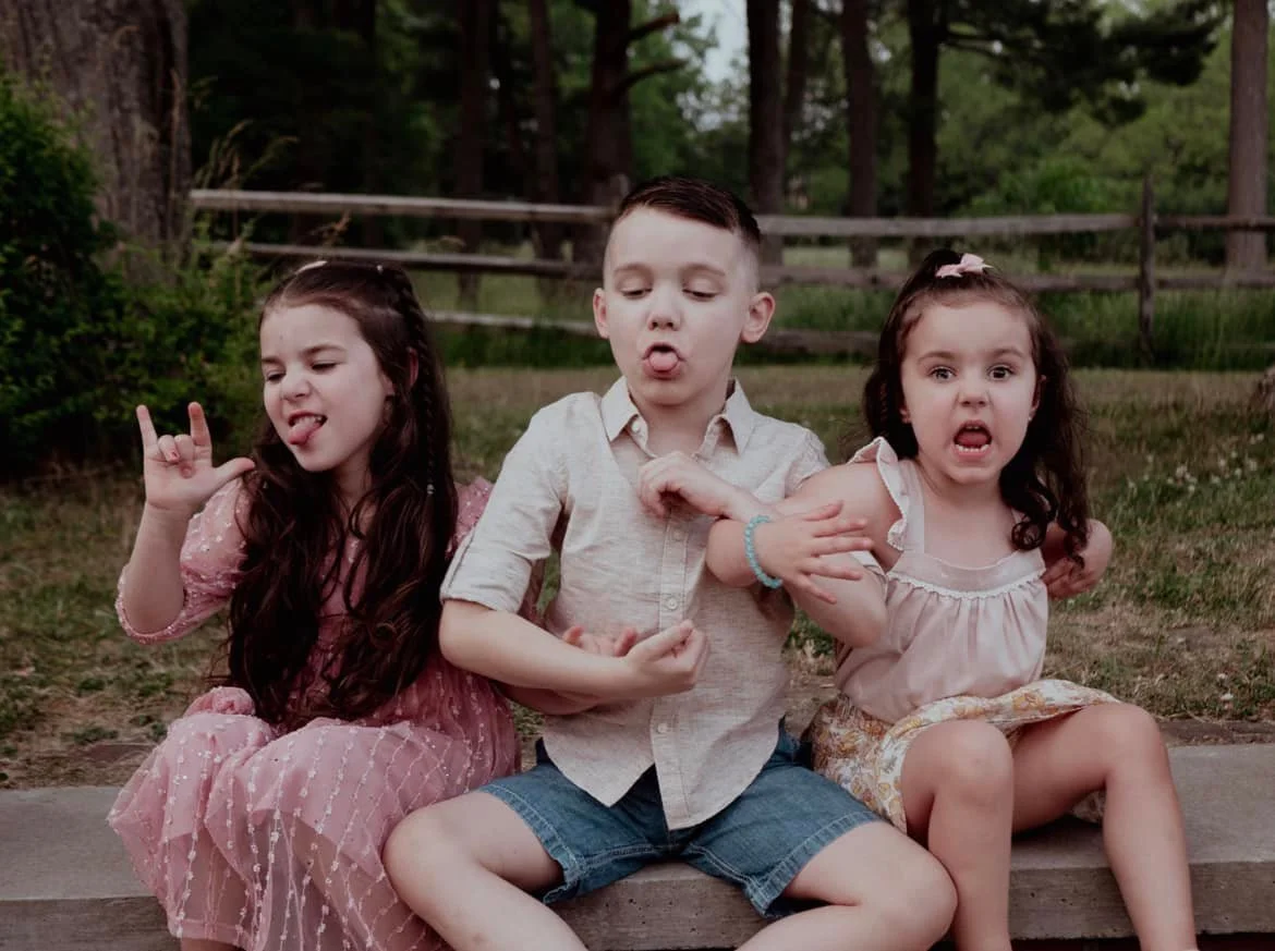 Three children sitting on a curb making silly faces and gestures in an outdoor park setting.