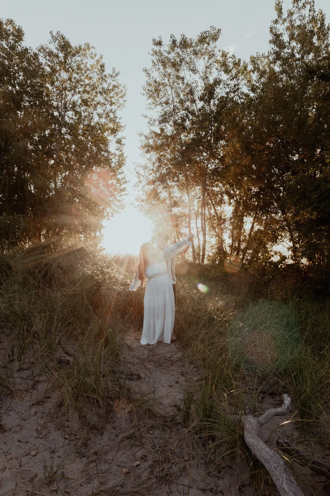 A woman in a white dress standing on a sandy path surrounded by tall grass and trees, with sunlight shining through the trees in the background.