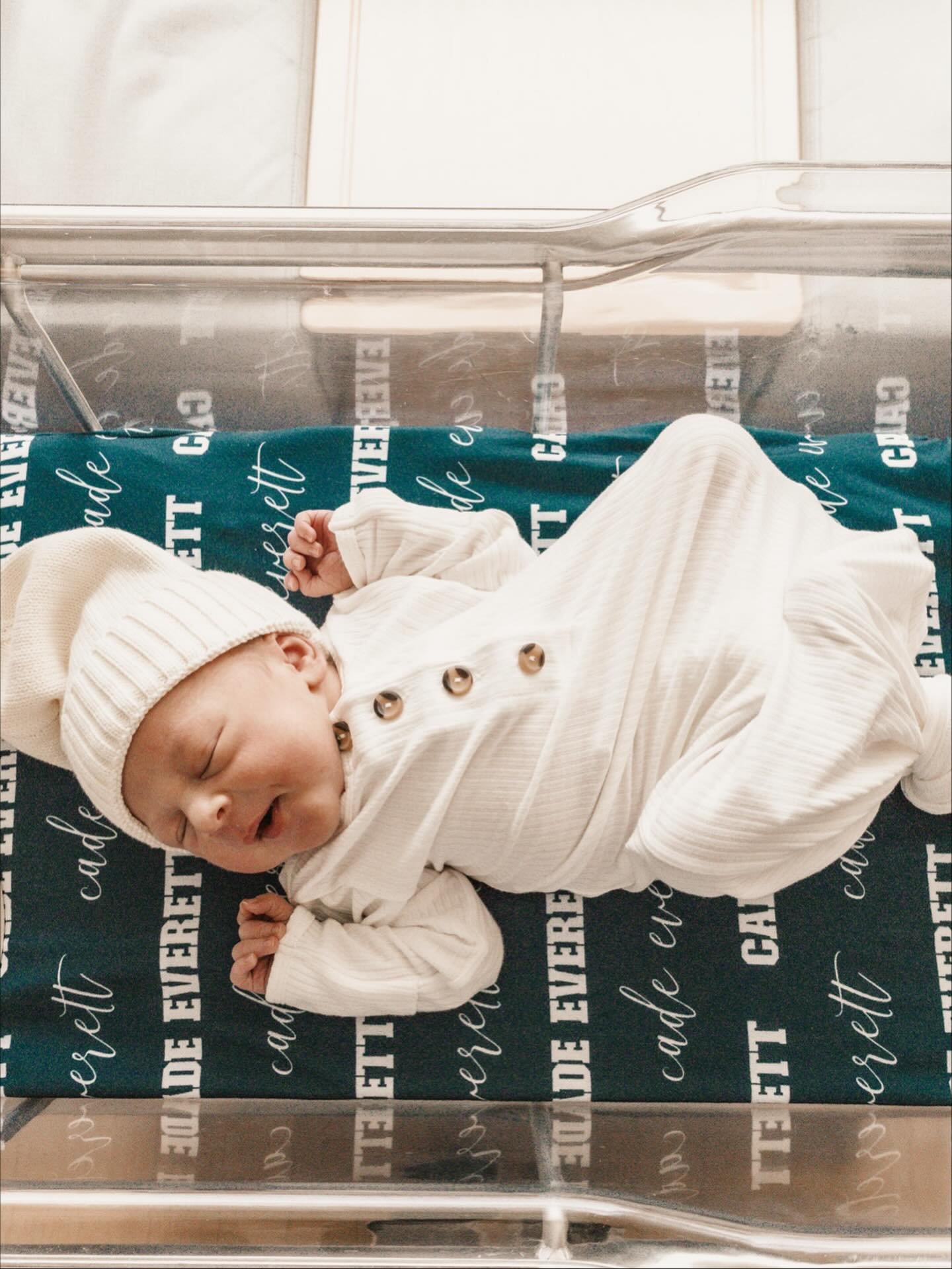 A newborn baby lying in a hospital bassinet, wearing a white knitted hat and a white outfit, smiling with eyes closed.