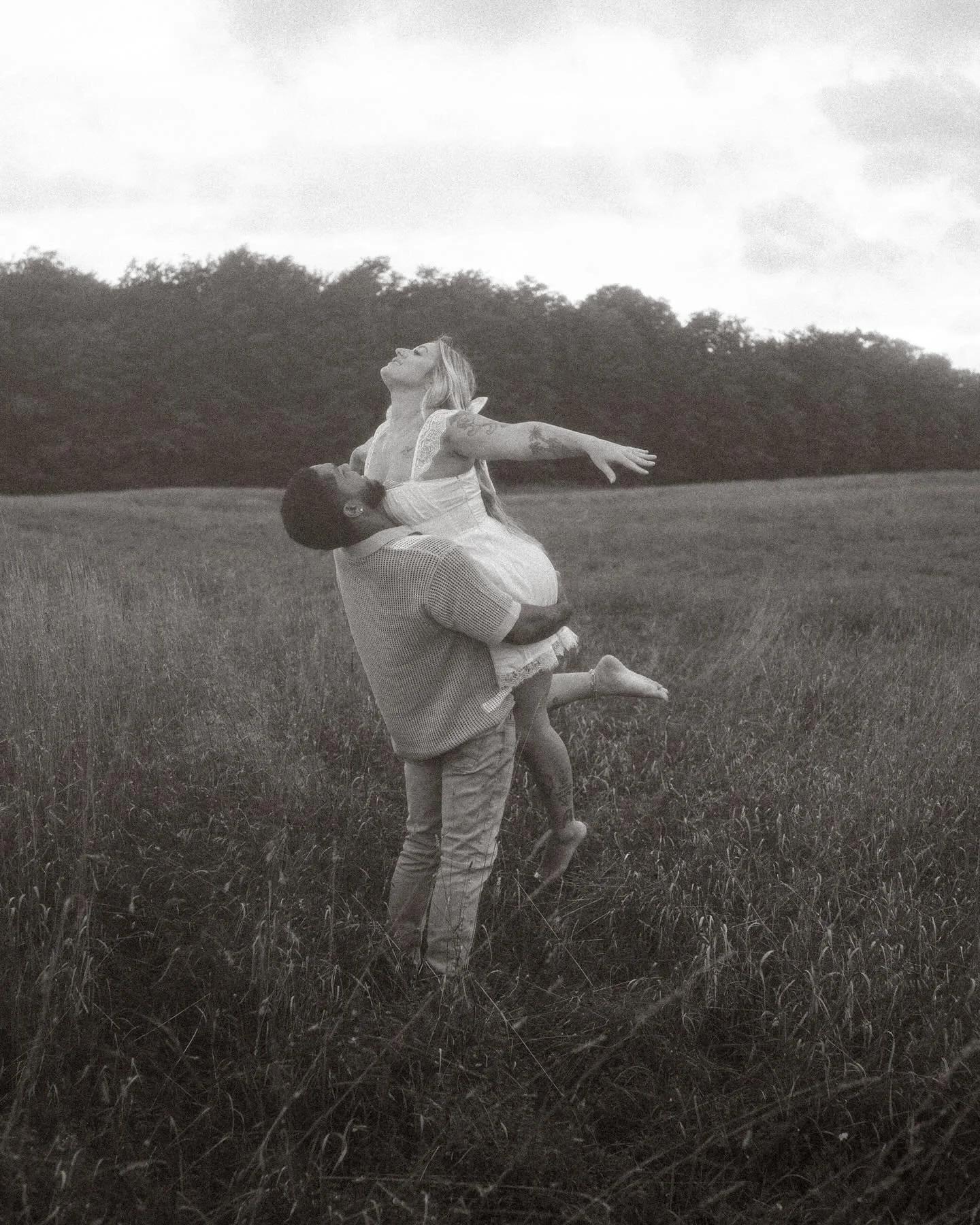 A man lifting a woman in a field, both smiling with open arms, black and white photograph.