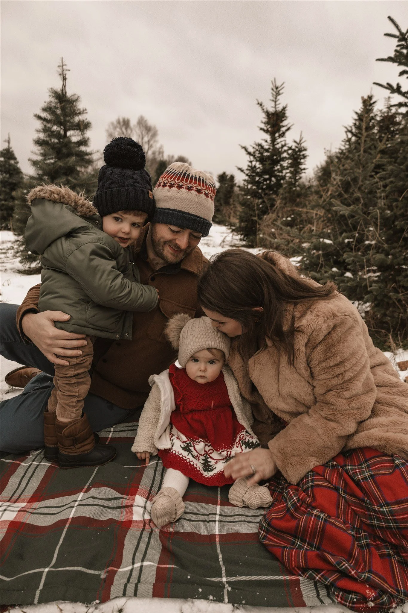 A family of four sitting on a plaid blanket outdoors in a snowy forest during winter, dressed in cozy winter clothing, enjoying each other's company.